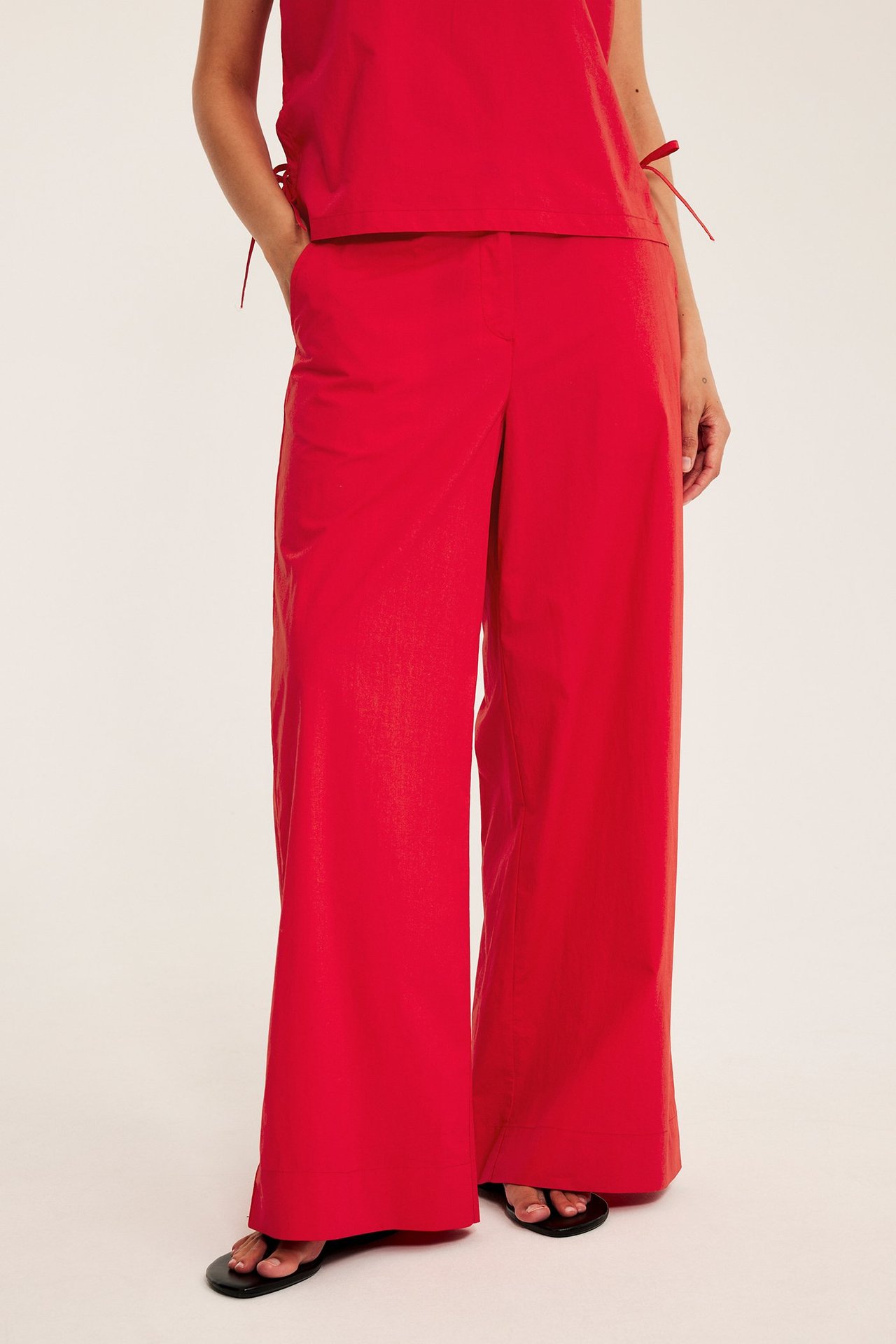 Framifrån: kvinna i röda vida byxor i bomullspoplin, matchande röd topp och svarta platta sandaler.