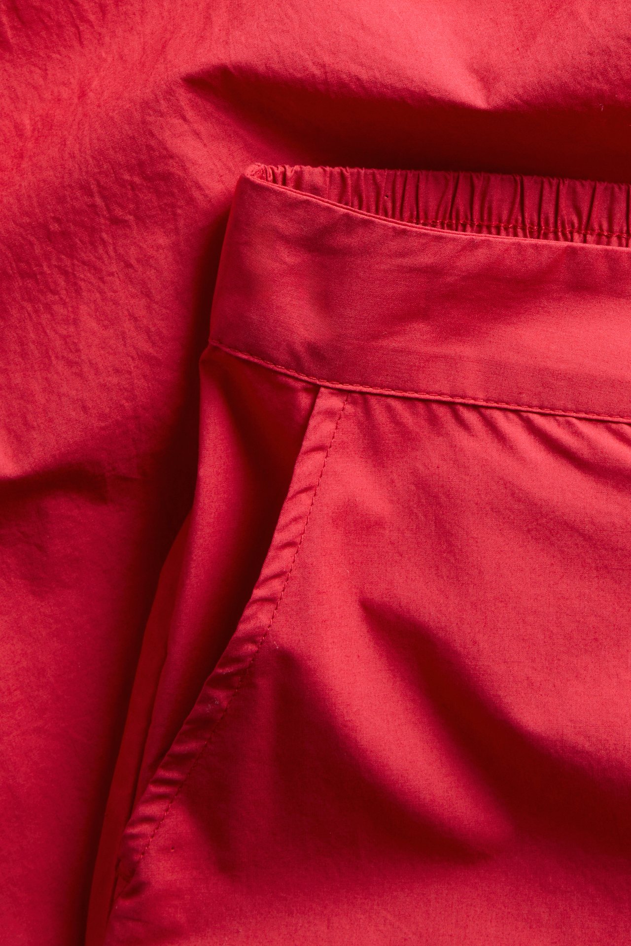Närbild av enfärgade röda byxor i bomullspoplin med sidoficka och elastisk midja baktill.