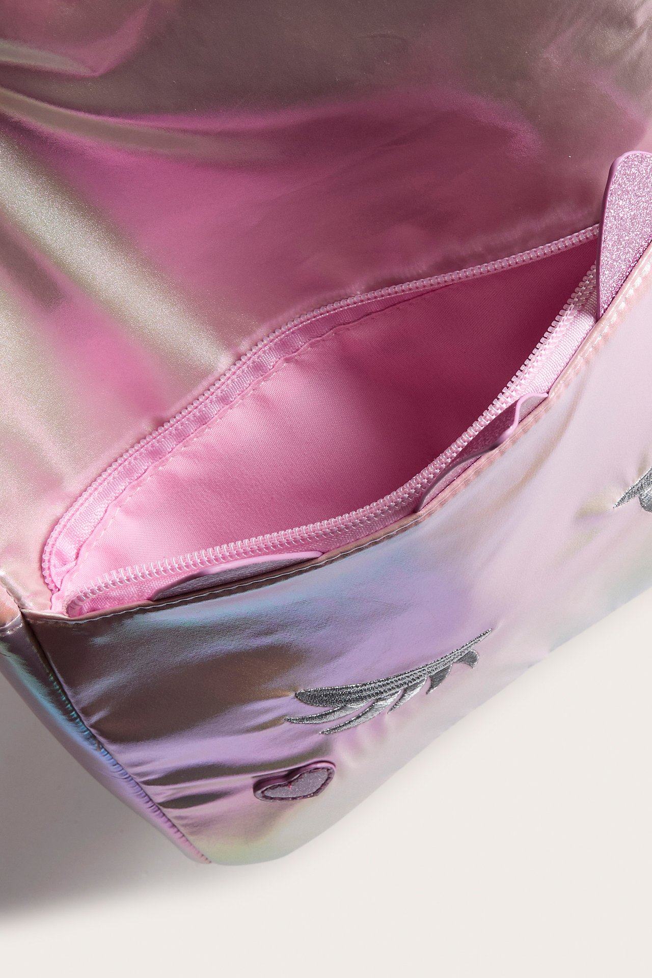 Lähikuva avoimesta irisoivasta laukusta, jossa on pinkki vuori, vetoketju, hopeakirjonta ja glitter-sydän.