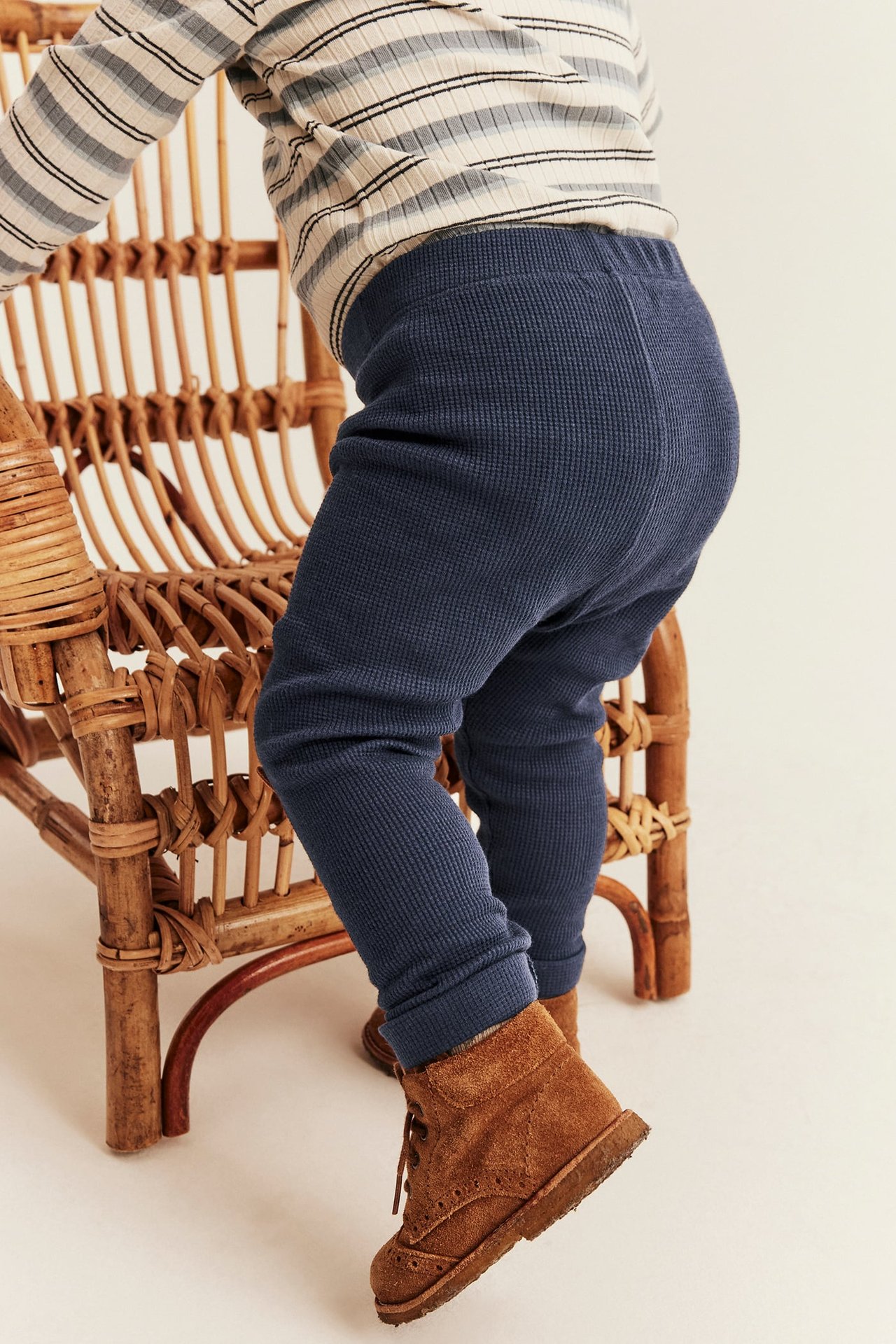 Sidorvy av bebis i marinblå våfflade leggings, en randig topp och bruna mockakängor.