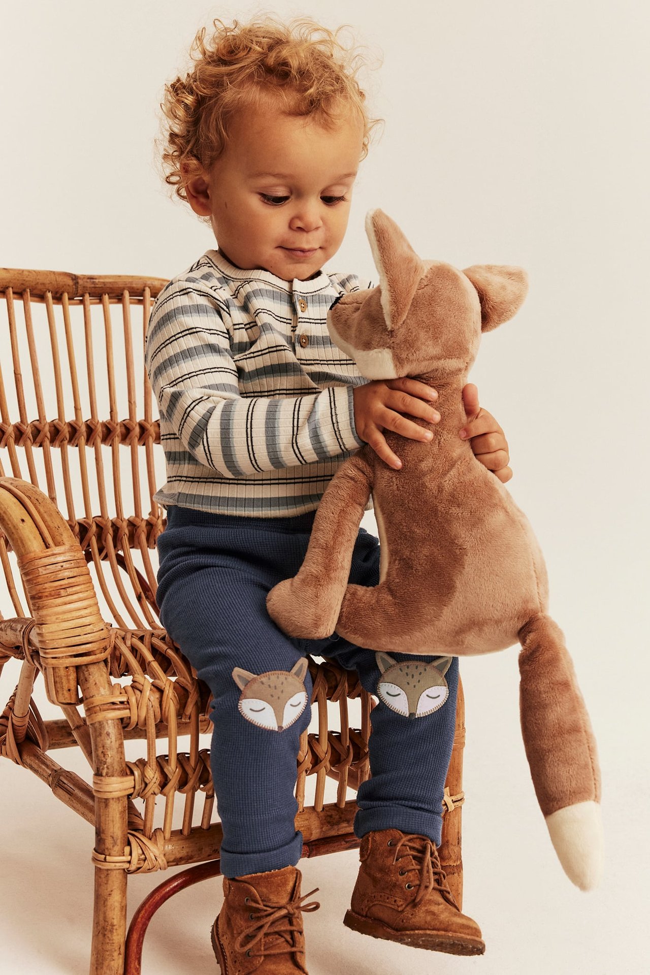 Frontbilde av baby i blå ribbestrikket leggings med reve-lapper, stripet topp og brune støvler, holder revebamse.