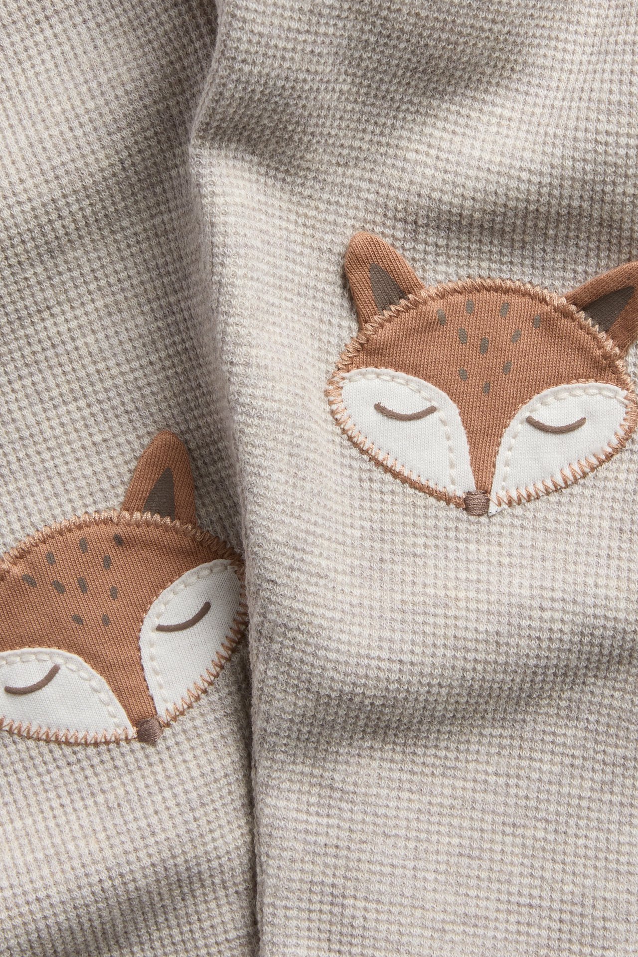 Närbild på ljust beigefärgat våfflat tyg med två broderade bruna rävansikten.