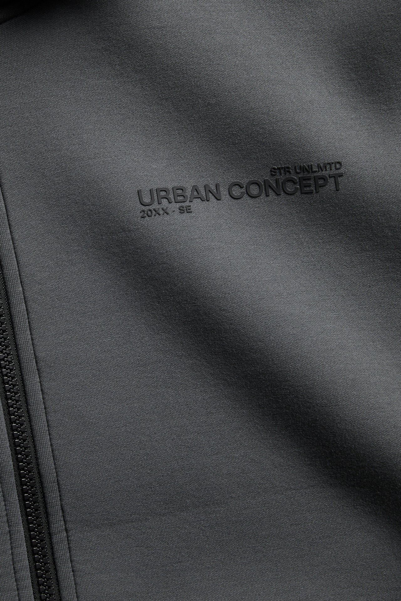 Närbild av en mörkgrå jacka med dragkedja och präglad text 'URBAN CONCEPT'.