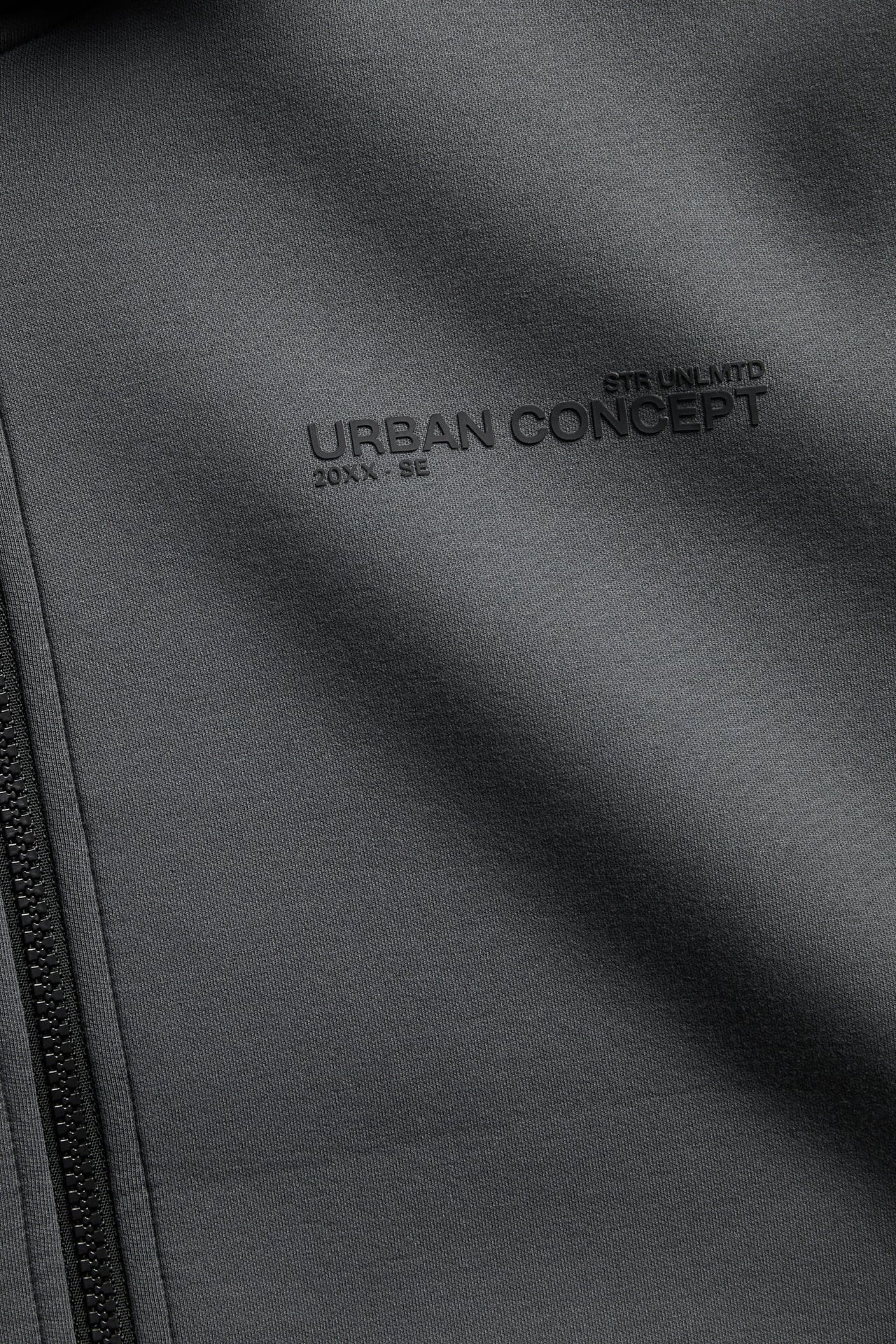Nærbilde av en mørkegrå jakke med glidelås og preget tekst 'URBAN CONCEPT'.