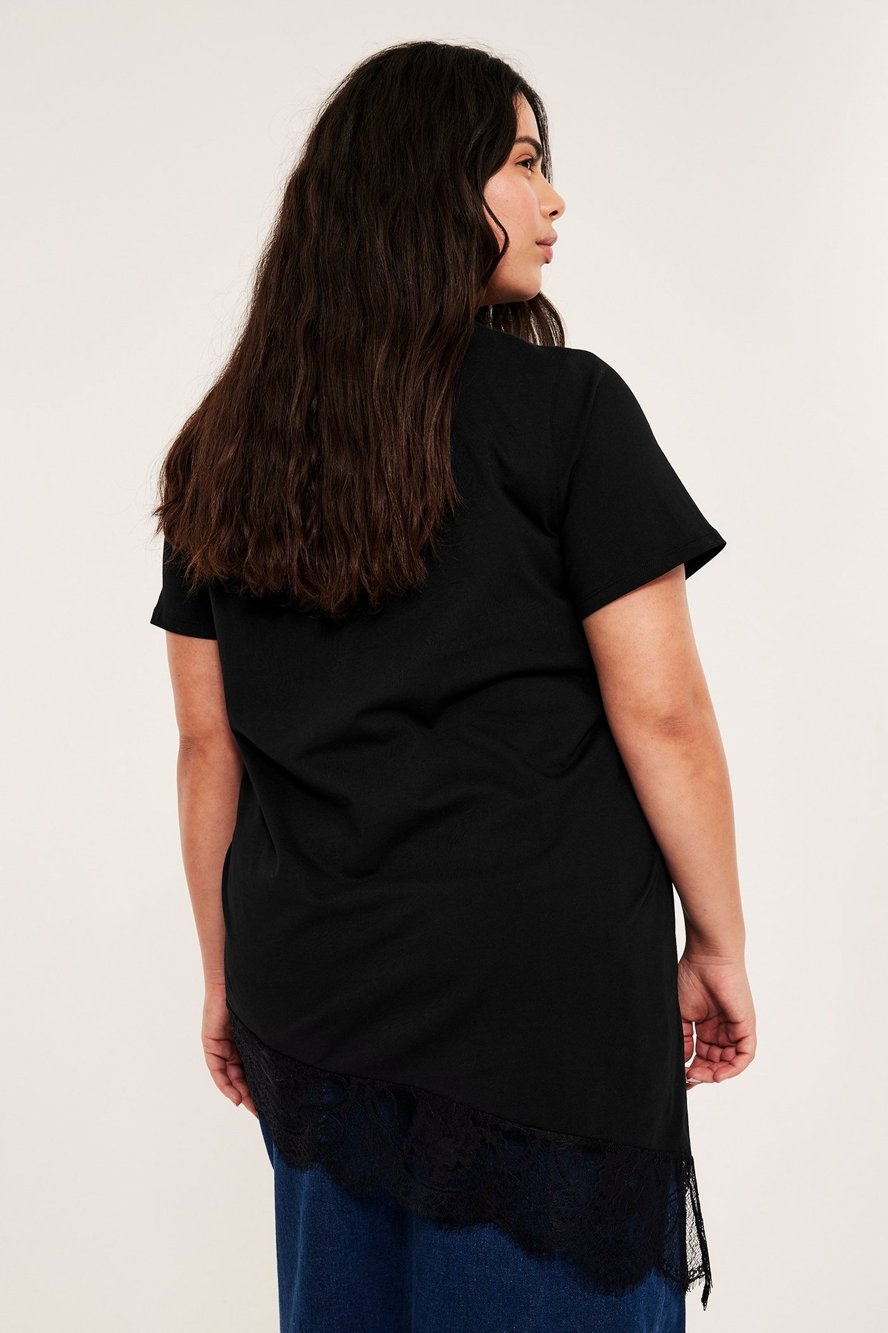 Bakfra av kvinne i svart kortermet asymmetrisk topp med blondekantet nederkant over blå jeans.