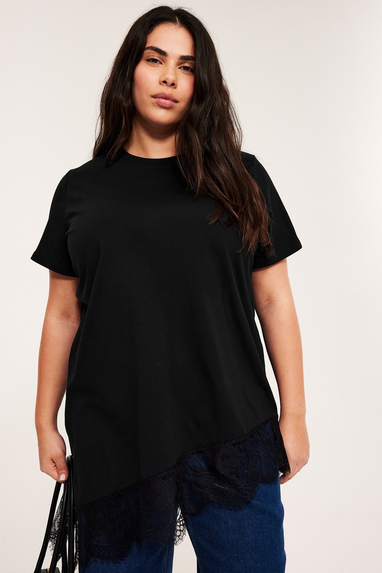 Framifrån: kvinna i svart kortärmad topp med asymmetrisk spetskant över blå jeans.