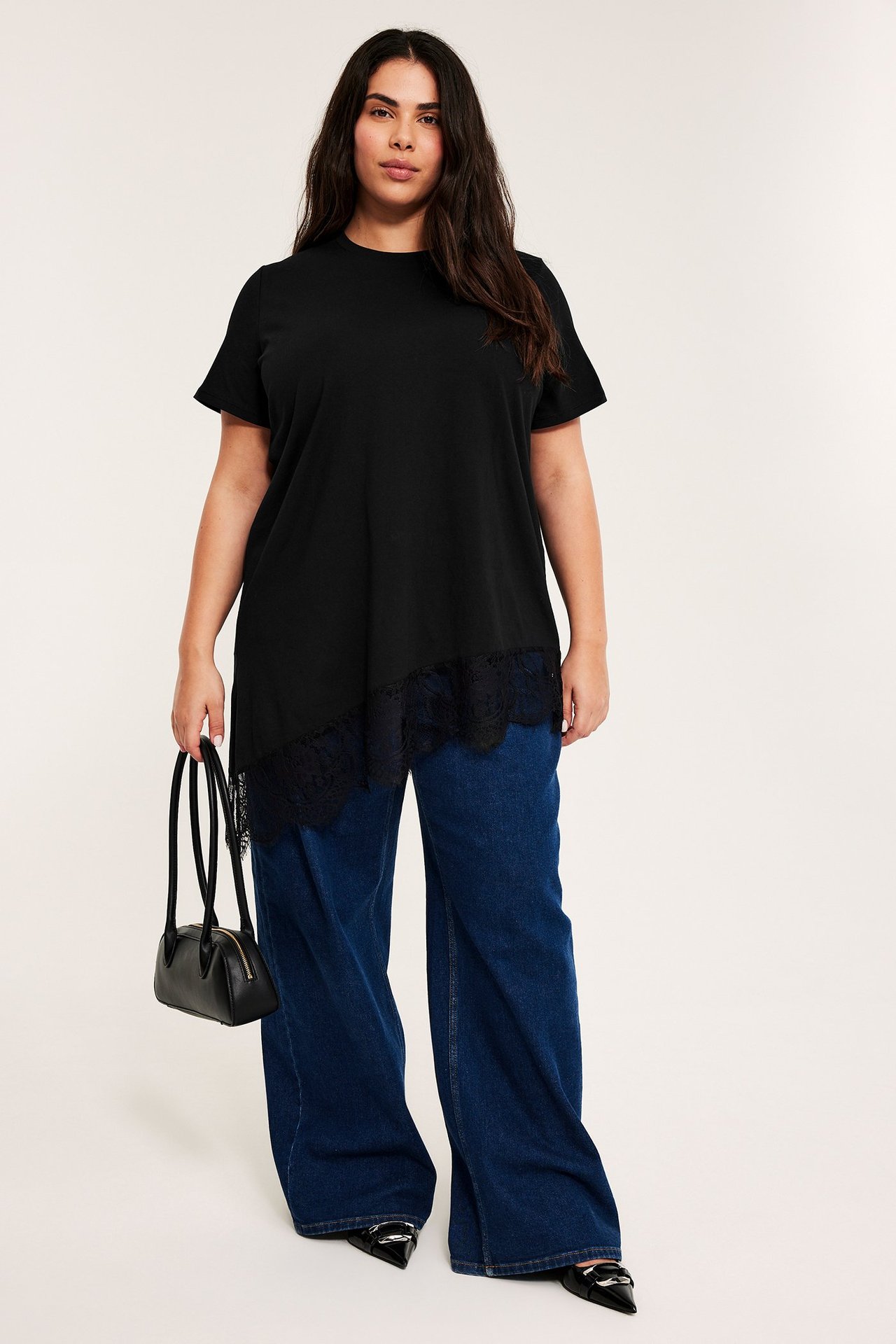 Framifrån, kvinna i svart asymmetrisk topp med spets, blå vida jeans, svarta spetsiga loafers, och håller en svart handväska.