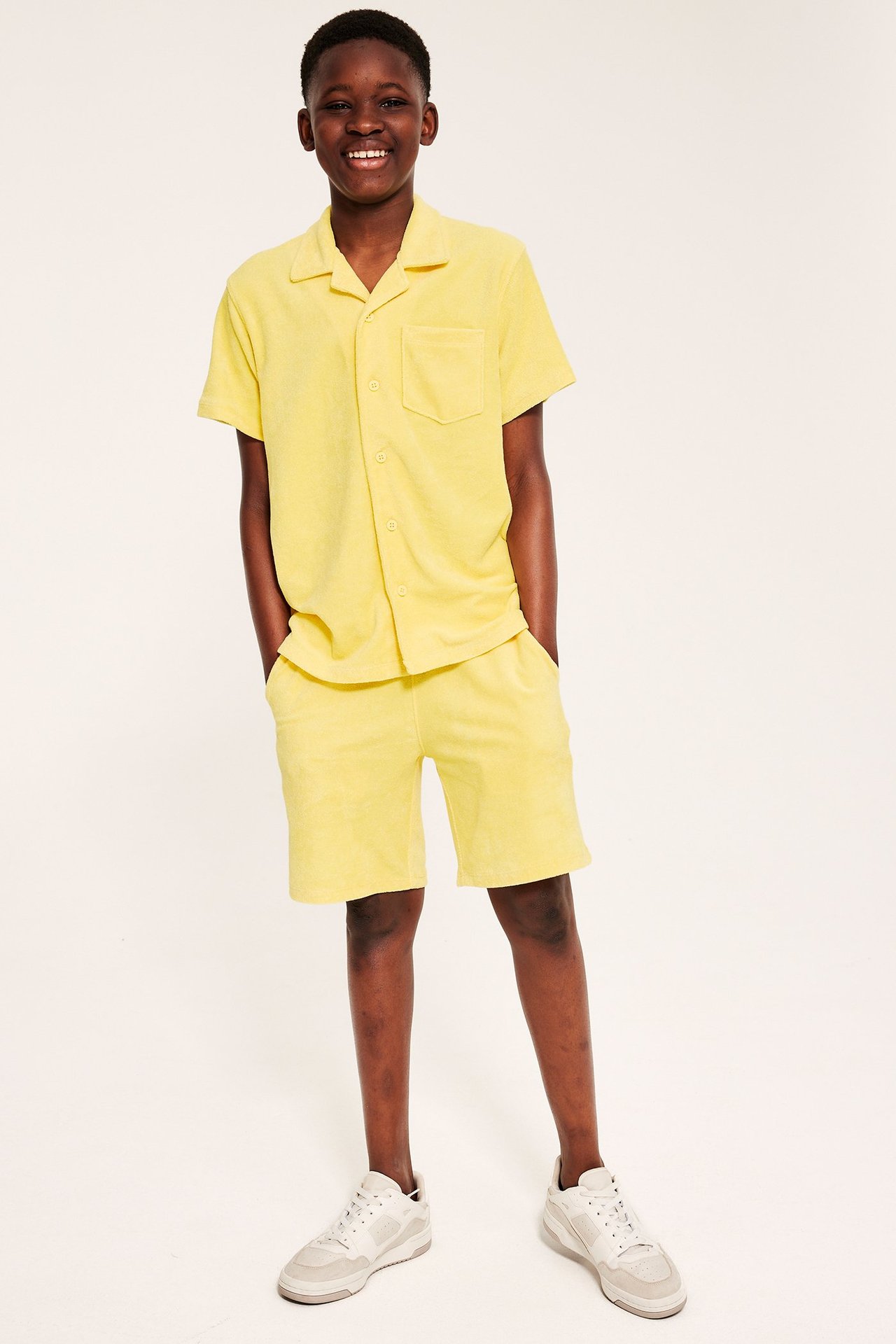 Edestäpäin hymyilevä poika keltaisissa froteeshortseissa, yhteensopivassa paidassa ja vaaleissa tennareissa.