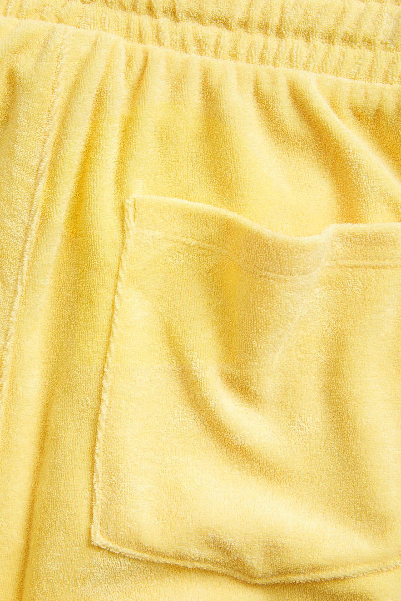 Närbild av gula frottéshorts med elastisk dragsko i midjan och en påsydd bakficka.