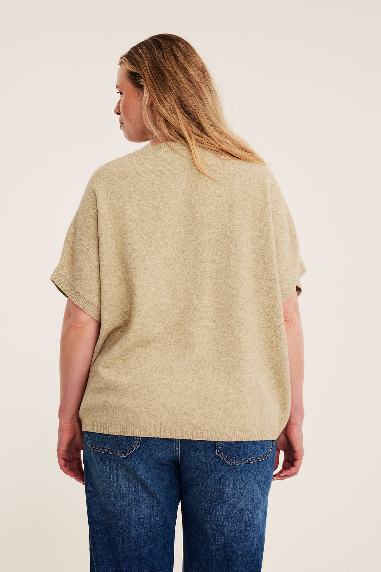 Bakfra kvinne i løs beige kortermet strikket topp og blå jeans.