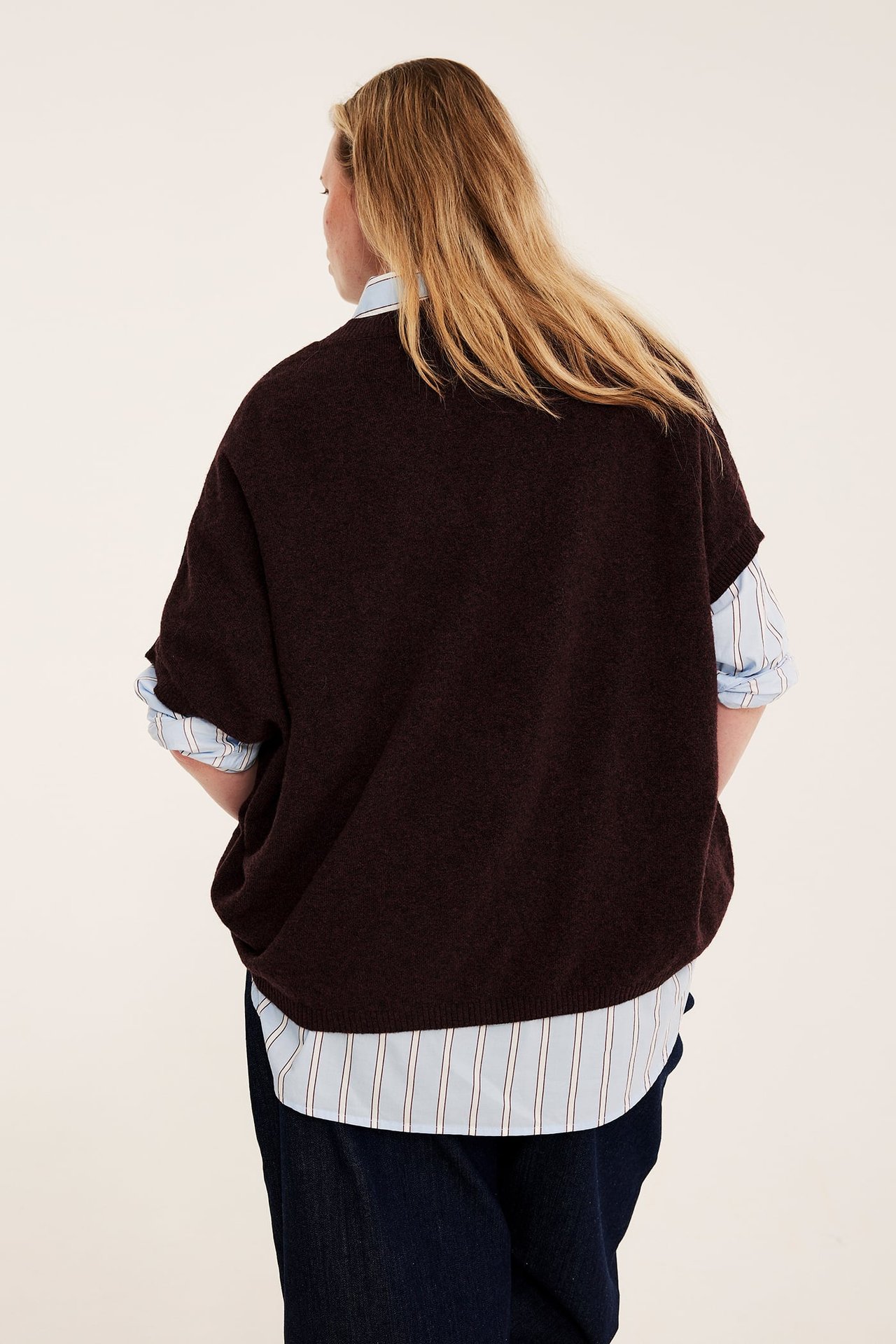Kvinna, bakifrån, i mörkbrun oversized tröja över randig skjorta och mörkblå jeans.