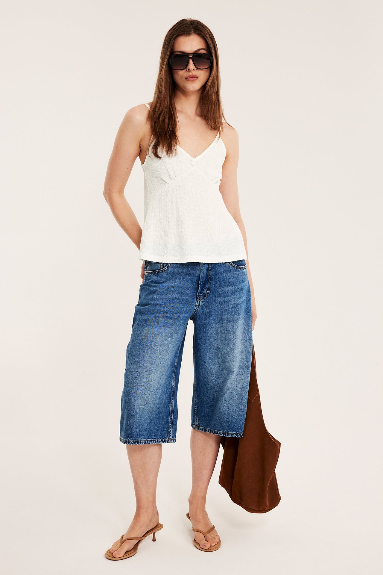 Frontvy av kvinna i offwhite strukturerat linne, blå denim culottes, bruna sandaler, svarta solglasögon, brun väska.