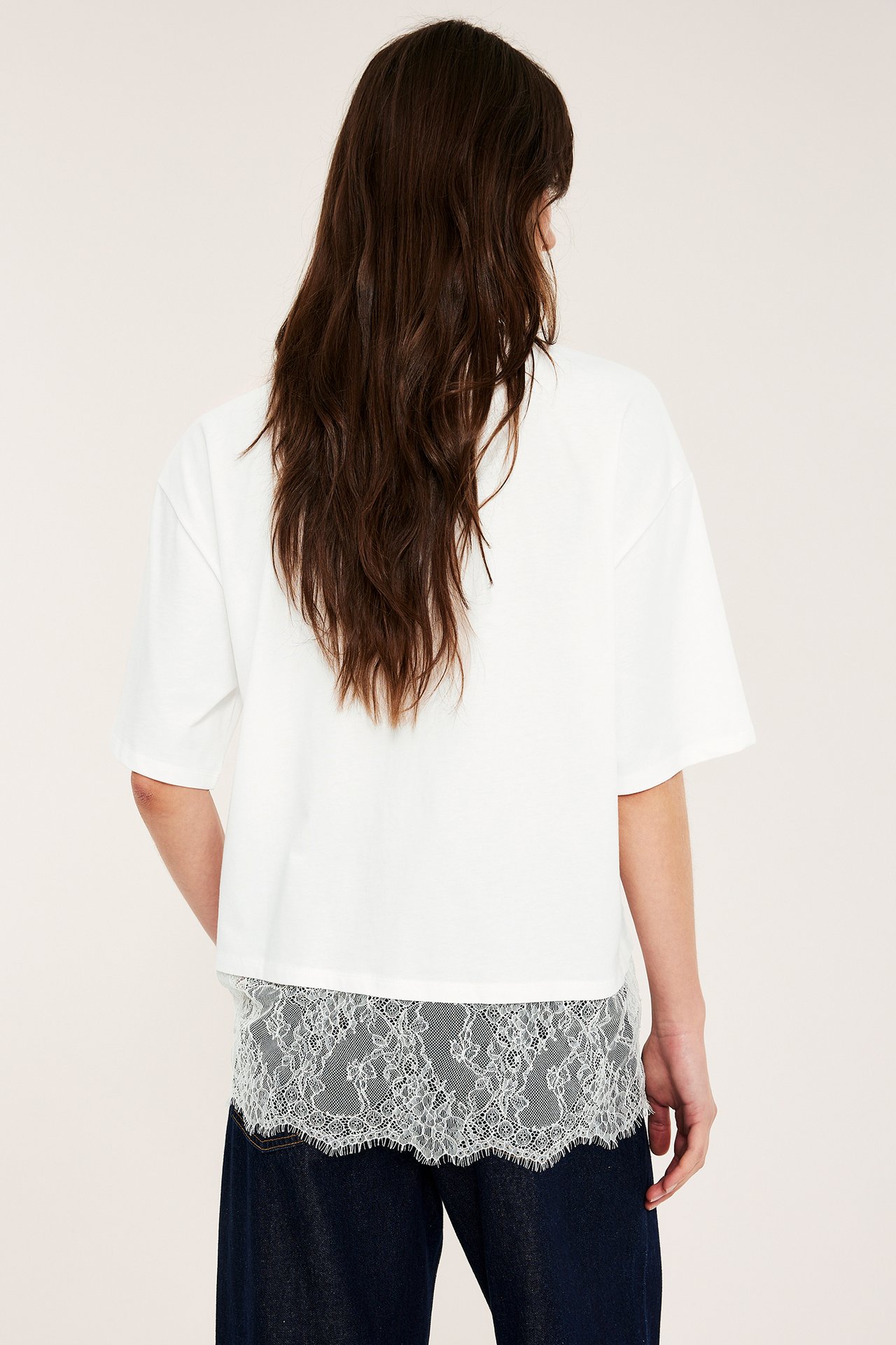 Bakfra: Kvinne i en offwhite løs t-skjorte med hvit blondekant over mørke jeans.