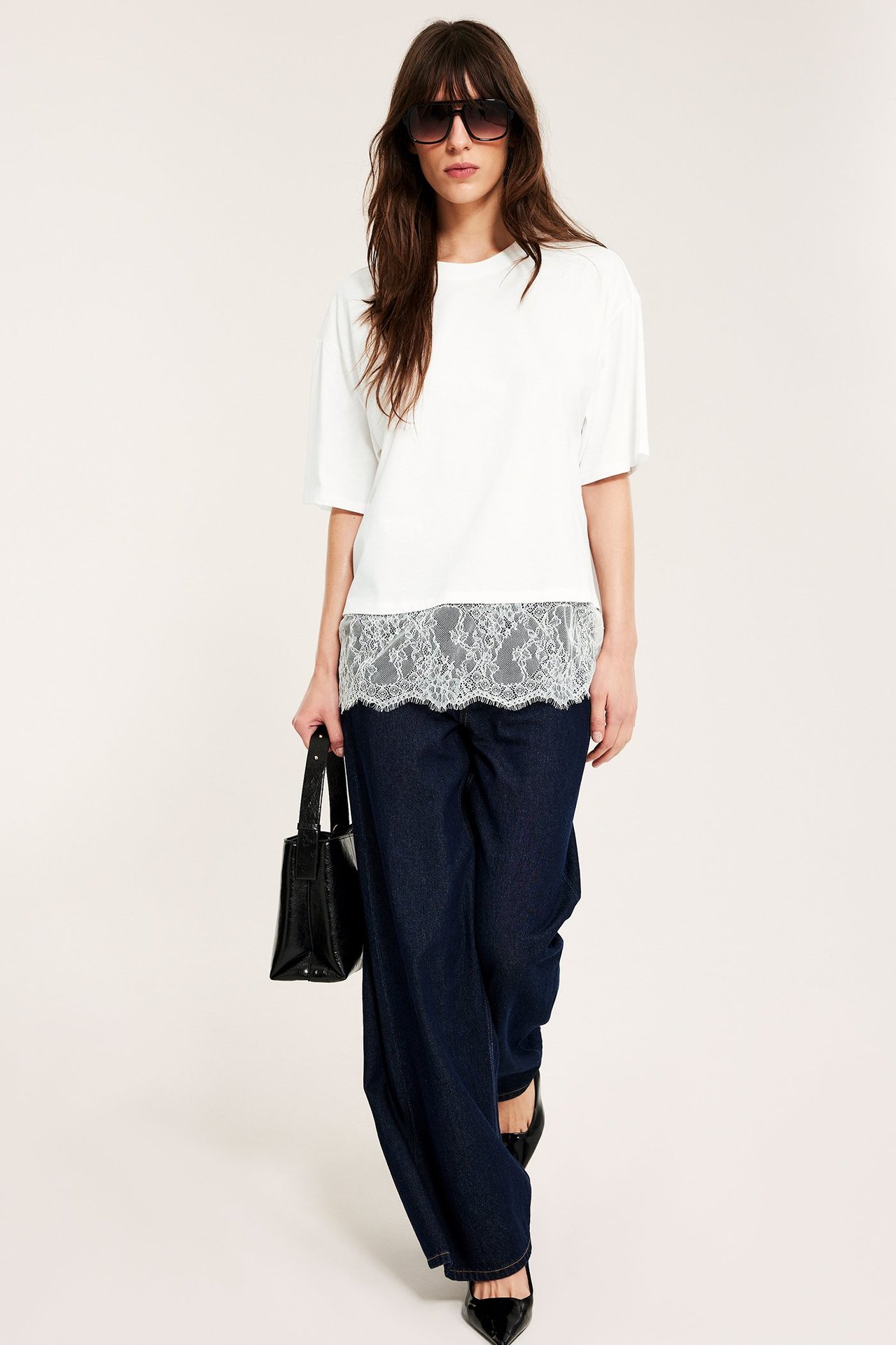 Widok z przodu: Kobieta w luźnej koszulce off-white z koronką, ciemnych jeansach, czarnych szpilkach, czarna torebka, okulary.