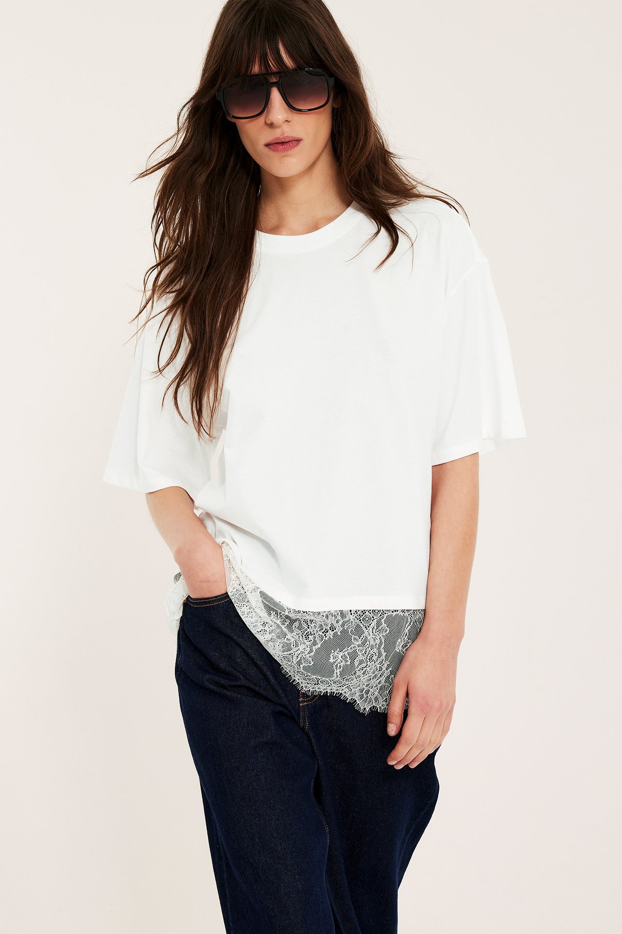 Frontvy: kvinna i offwhite lös t-shirt med spetskant, mörkblå jeans och solglasögon.