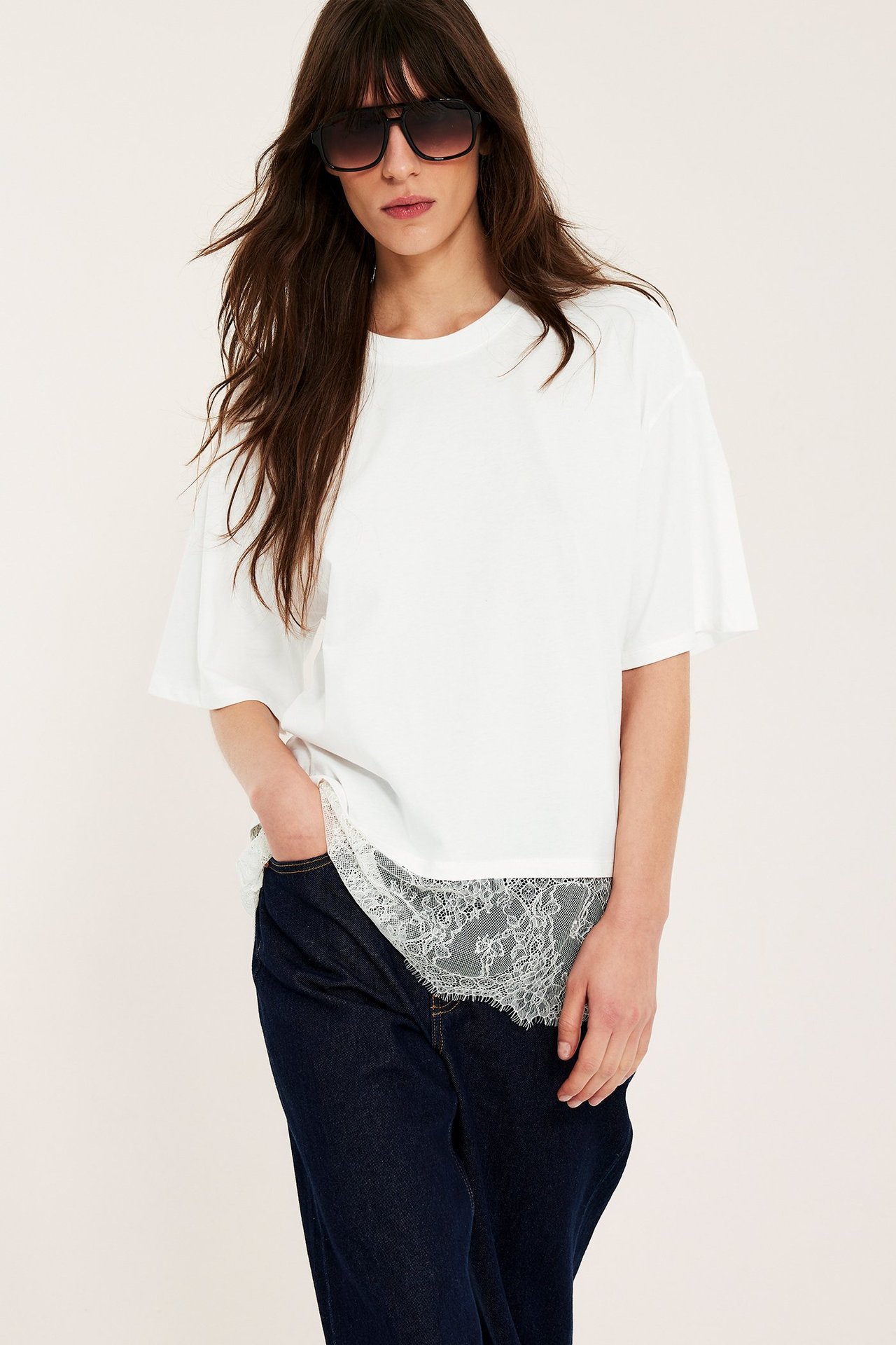 Widok z przodu: kobieta w luźnym T-shircie ecru z koronkowym dołem, ciemnych dżinsach i okularach.