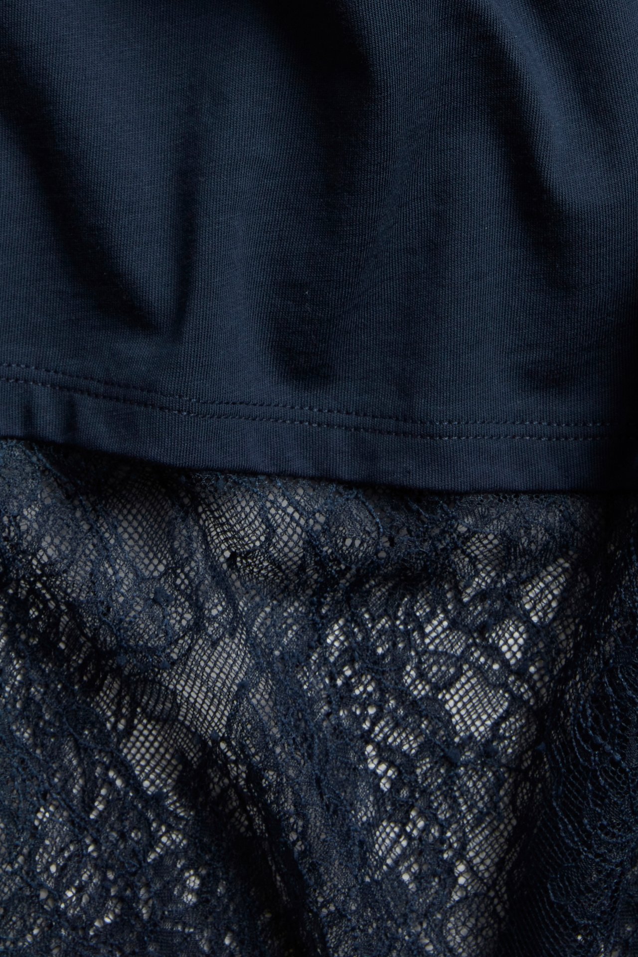 Lähikuva tummansinisestä t-paidasta, jossa on yksivärinen yläosa ja läpikuultava pitsireuna helmassa.