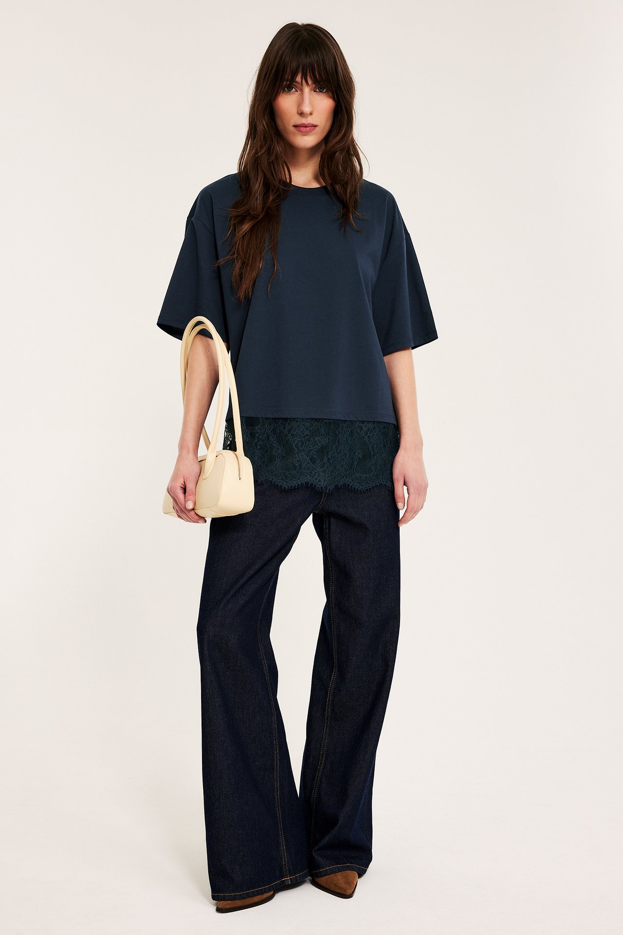 Framifrån: Kvinna i mörkblå lös t-shirt med grön spetskant, mörkblå jeans och hållande beige handväska.