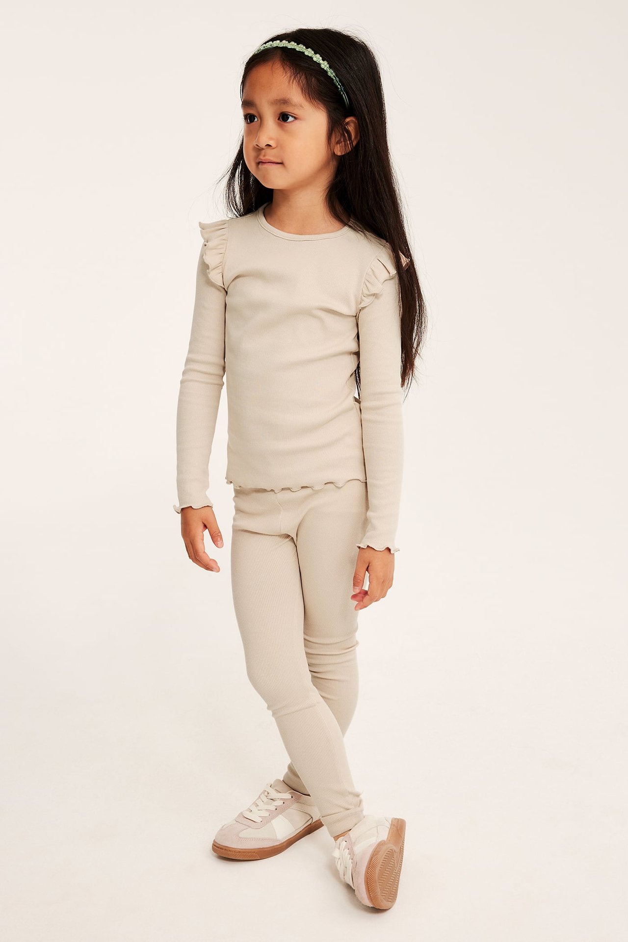Sidovy av ett barn i ljusbeige ribbad långärmad topp med volanger, matchande leggings, sneakers, grönt hårband.