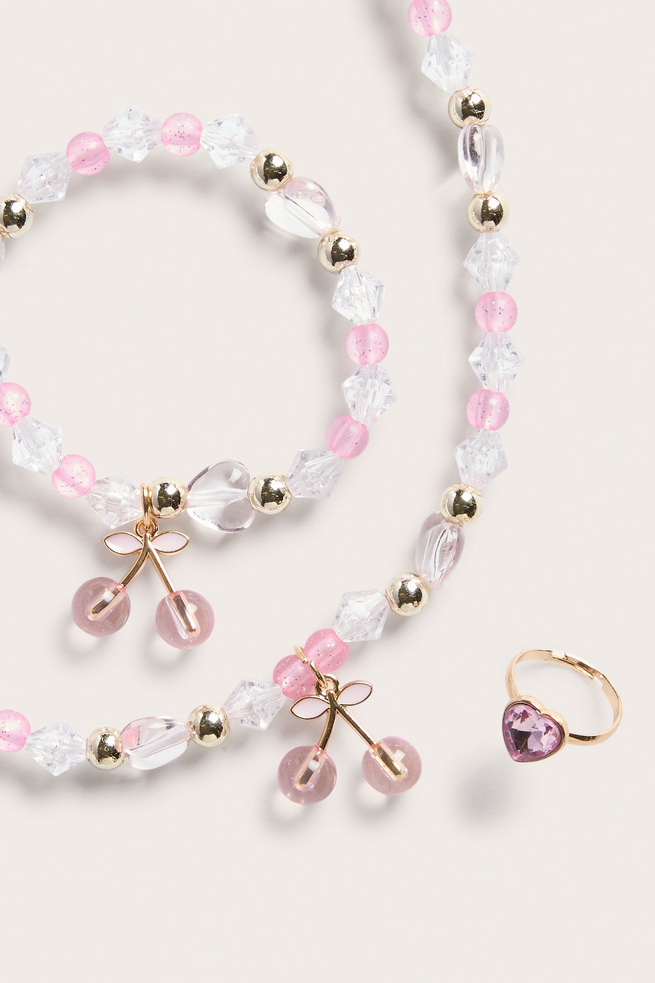 Närbild av ett rosa, transparent och guldfärgat pärlhalsband och armband med körsbärsberlocker, plus en rosa hjärtring.