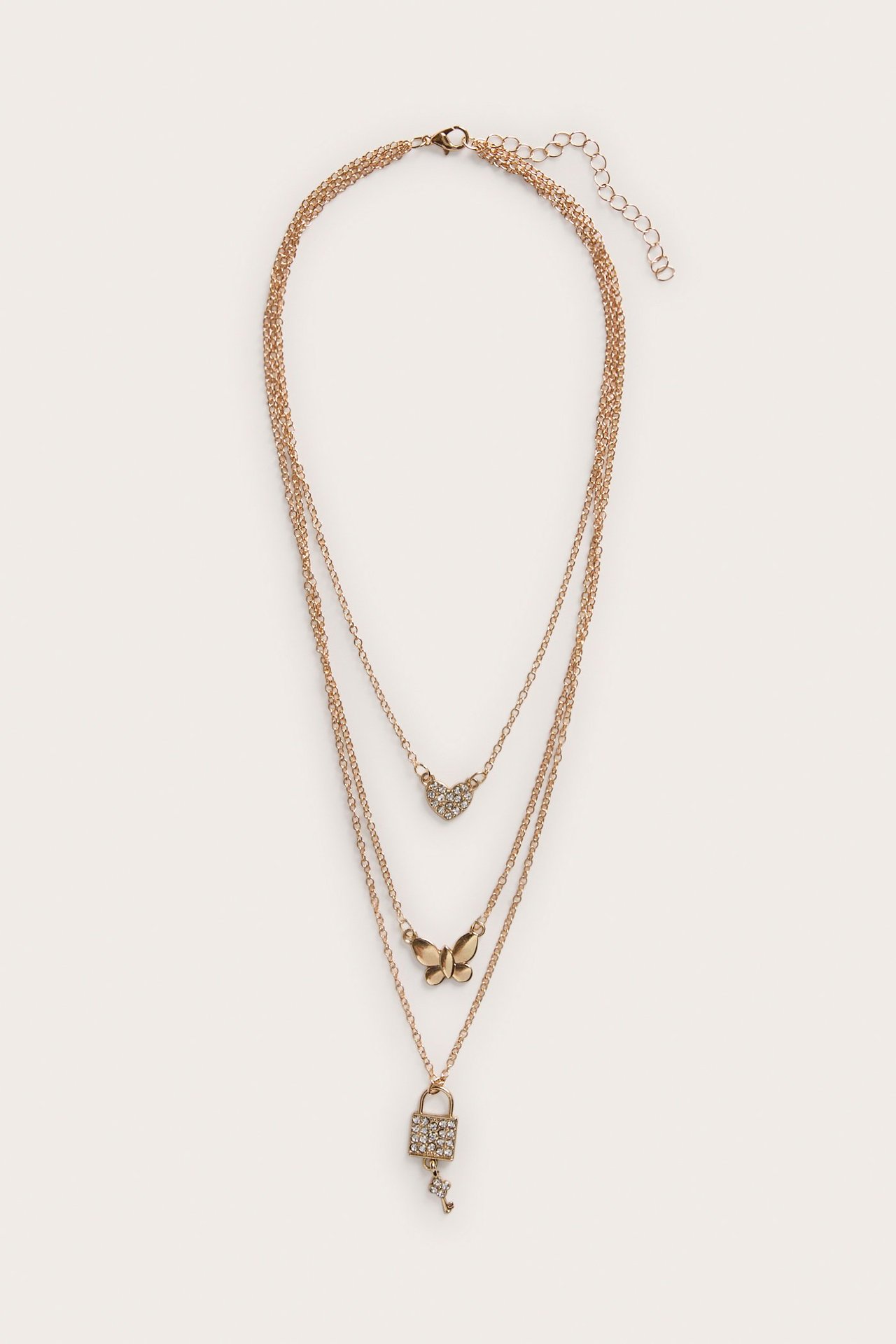 Etunäkymä kultaisesta kolmikerroksisesta kaulakorusta, jossa on strassikoristeiset sydän-, perhos-, ja lukko-avaimen riipukset.