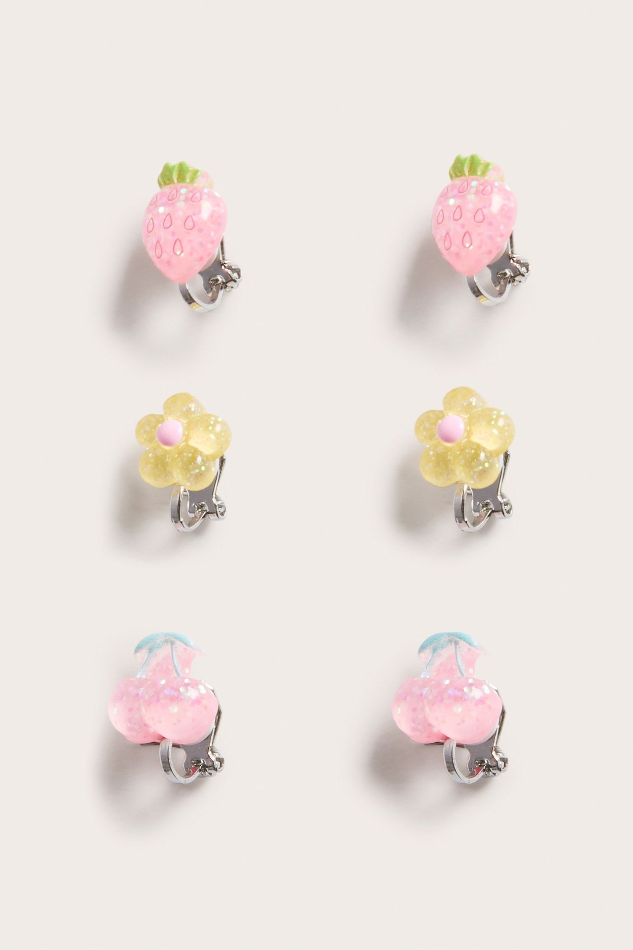Närbild på tre par glitteriga barnörhängen med clips: rosa jordgubbar, gula blommor och rosa körsbär.