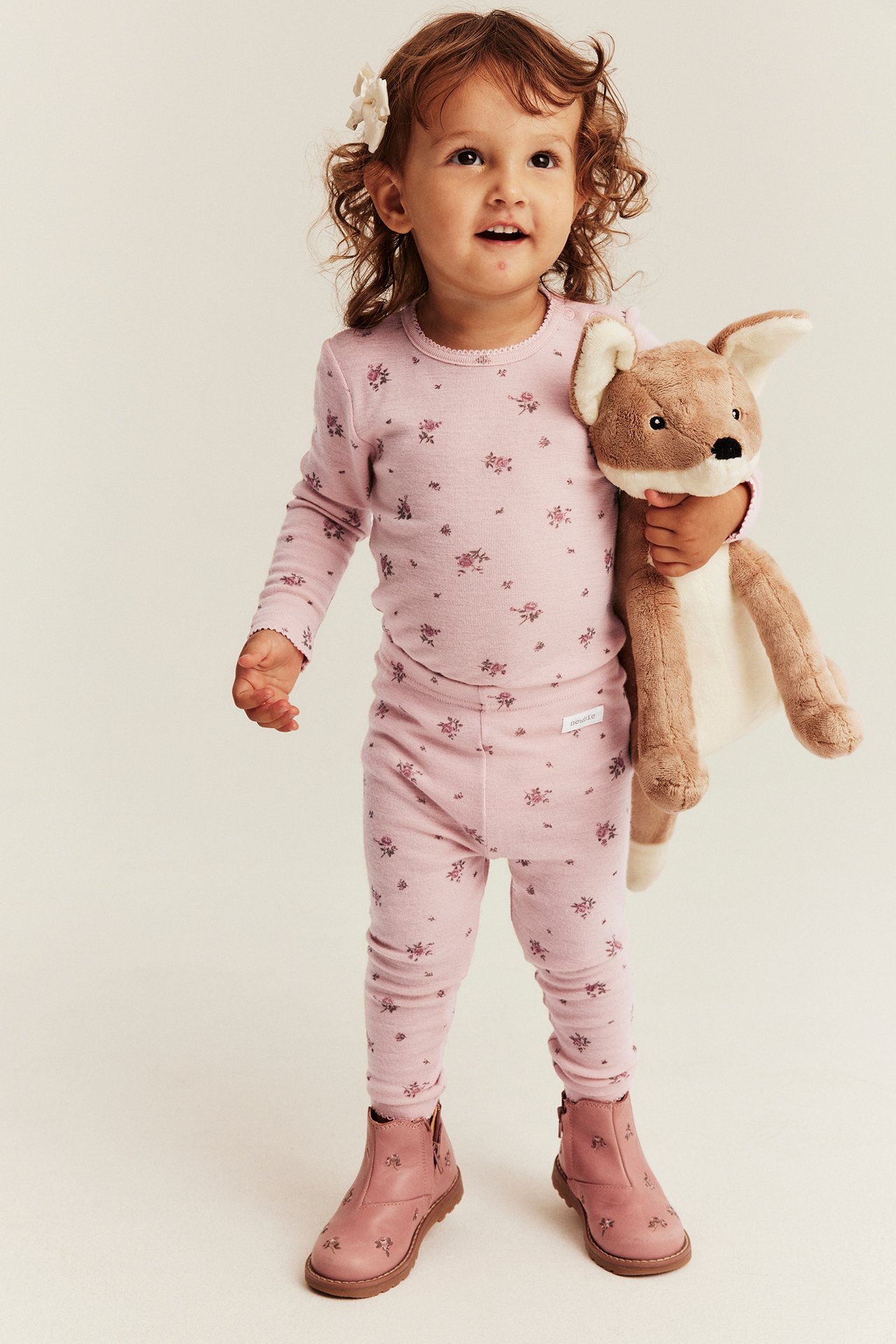 Edestä: lapsi yllään vaaleanpunainen kukkakuvioinen pitkähihainen pyjama, pinkit saappaat ja ruskea pehmolelu.