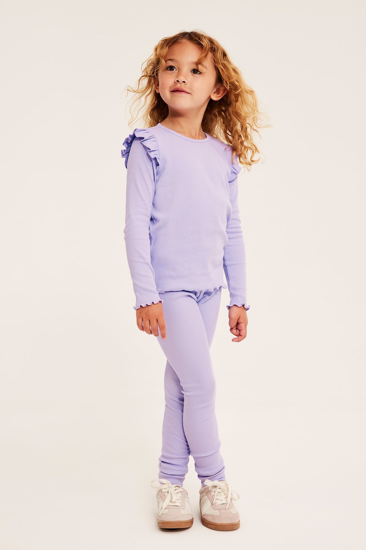 Frontbilde: Barn bruker en lilla langermet topp med rysjer, matchende leggings og beige sneakers.