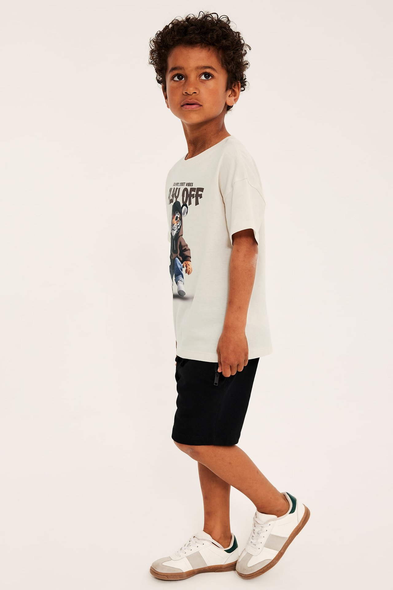 Sidovy av ett barn i en ljusbeige oversize bomulls-t-shirt med tigertryck, svarta shorts och vita sneakers.