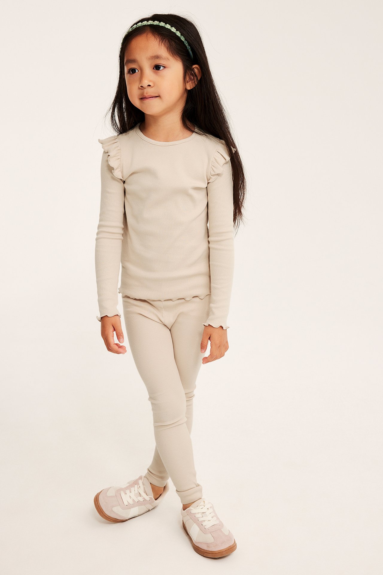 Forfra: et barn i lys beige ribbet bomullsleggings, matchende topp, grønt hårband og beige-rosa sneakers.
