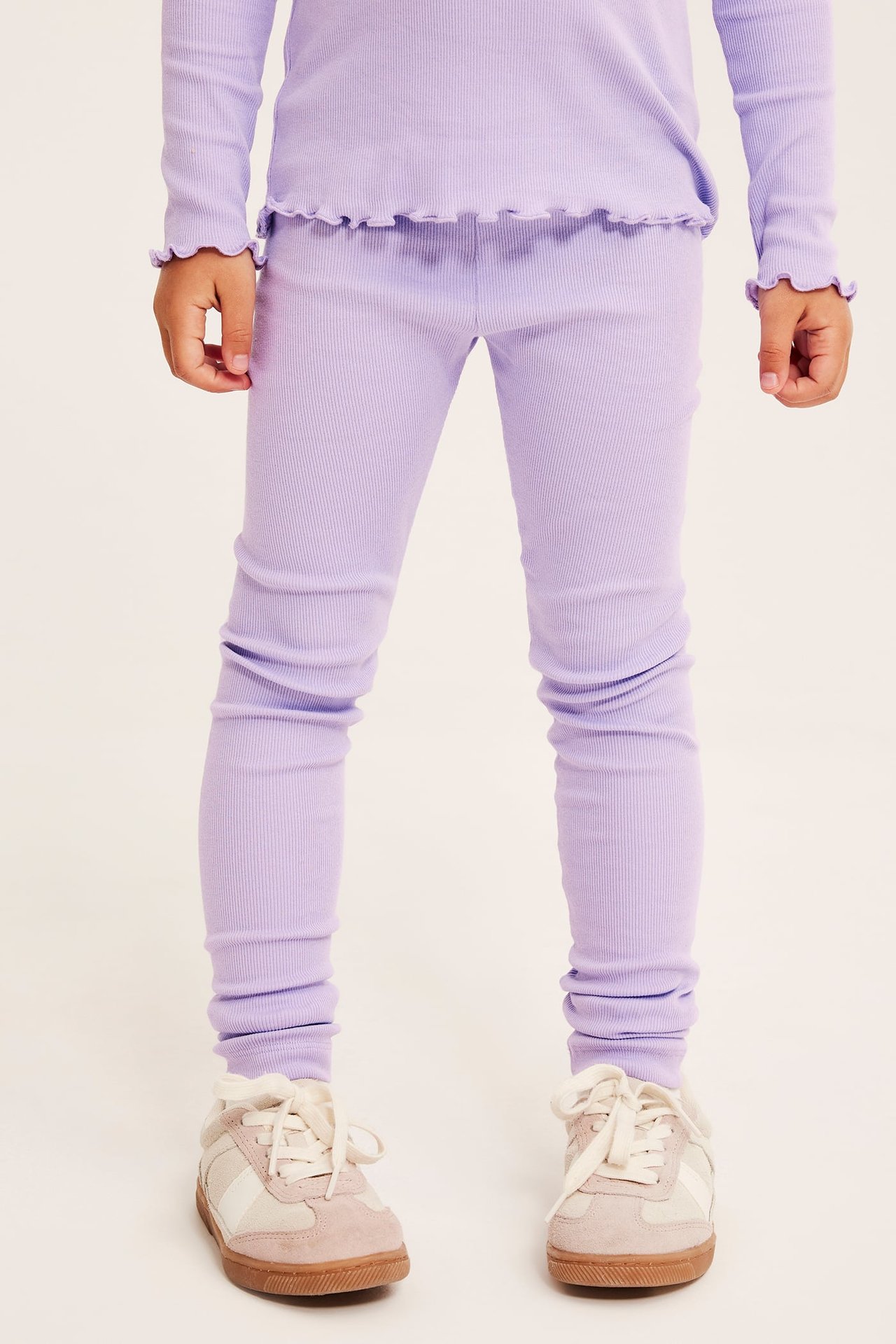 Edestä kuvattu lapsi yllään violetit ribbileggingsit, yhteensopiva röyhelötoppi ja beiget lenkkarit.