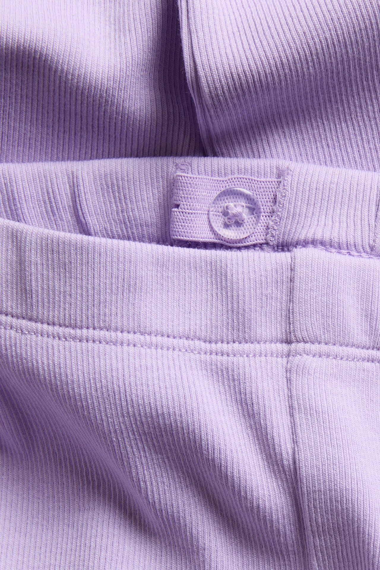 Lähikuva lasten vaaleanliloista ribbineulotuista puuvillaleggingsistä, joissa säädettävä joustovyötärö ja läpinäkyvä nappi.