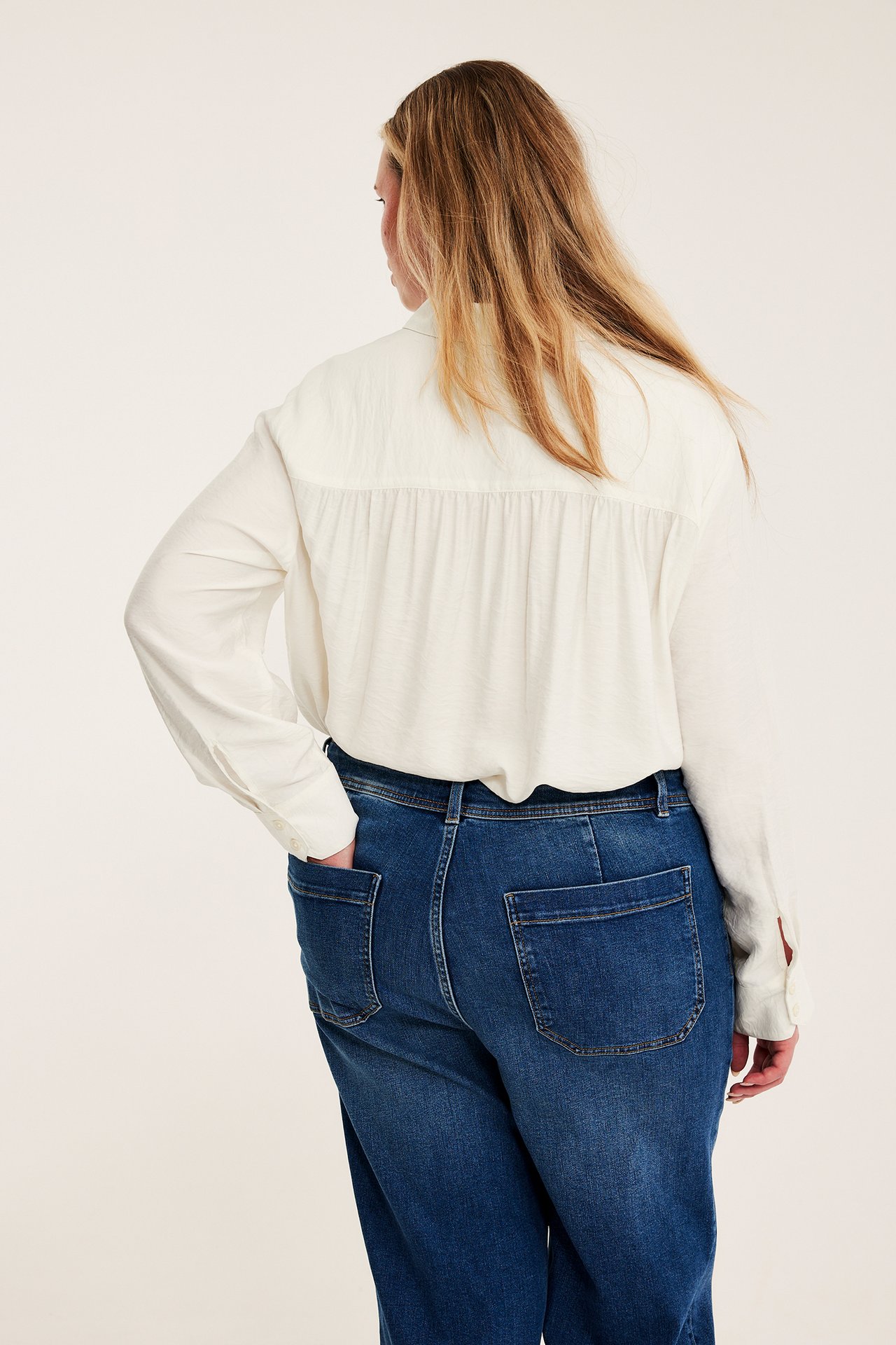 Kvinne, bakfra, i en hvit langermet skjorte med plissert rygg og blå jeans.