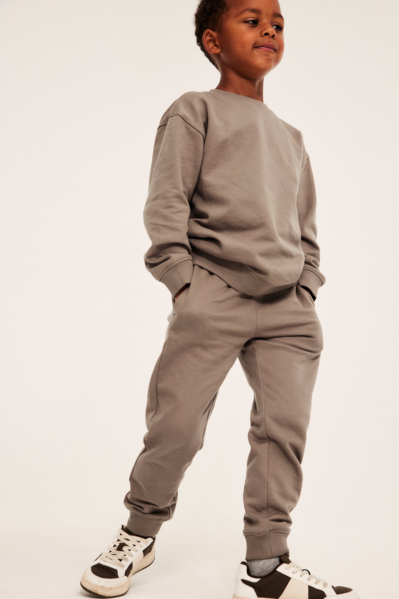 Framifrån, ett barn i en avslappnad taupe sweatshirt och matchande mjukisbyxor, med tvåfärgade sneakers.