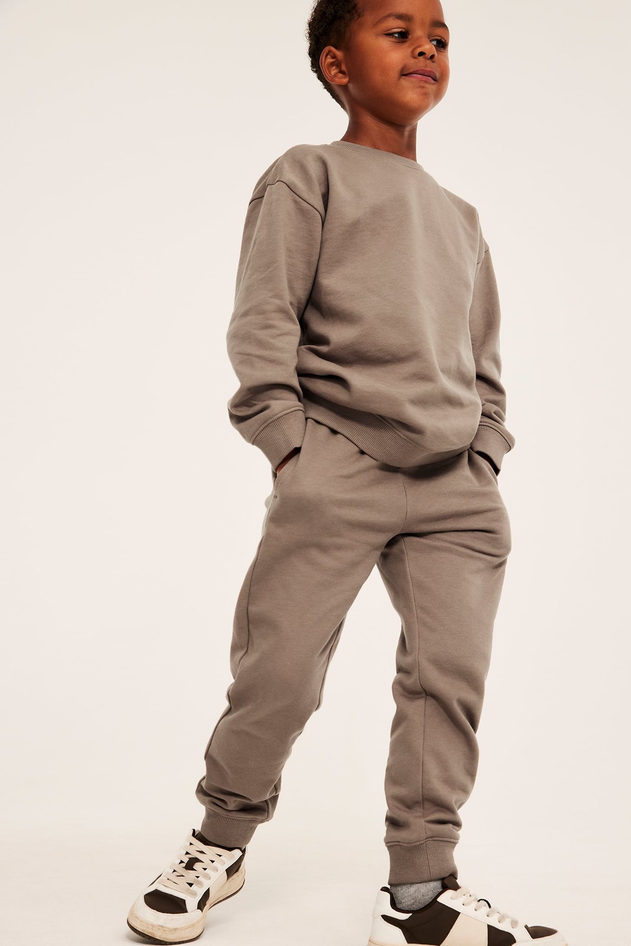 Framifrån, ett barn i en avslappnad taupe sweatshirt och matchande mjukisbyxor, med tvåfärgade sneakers.