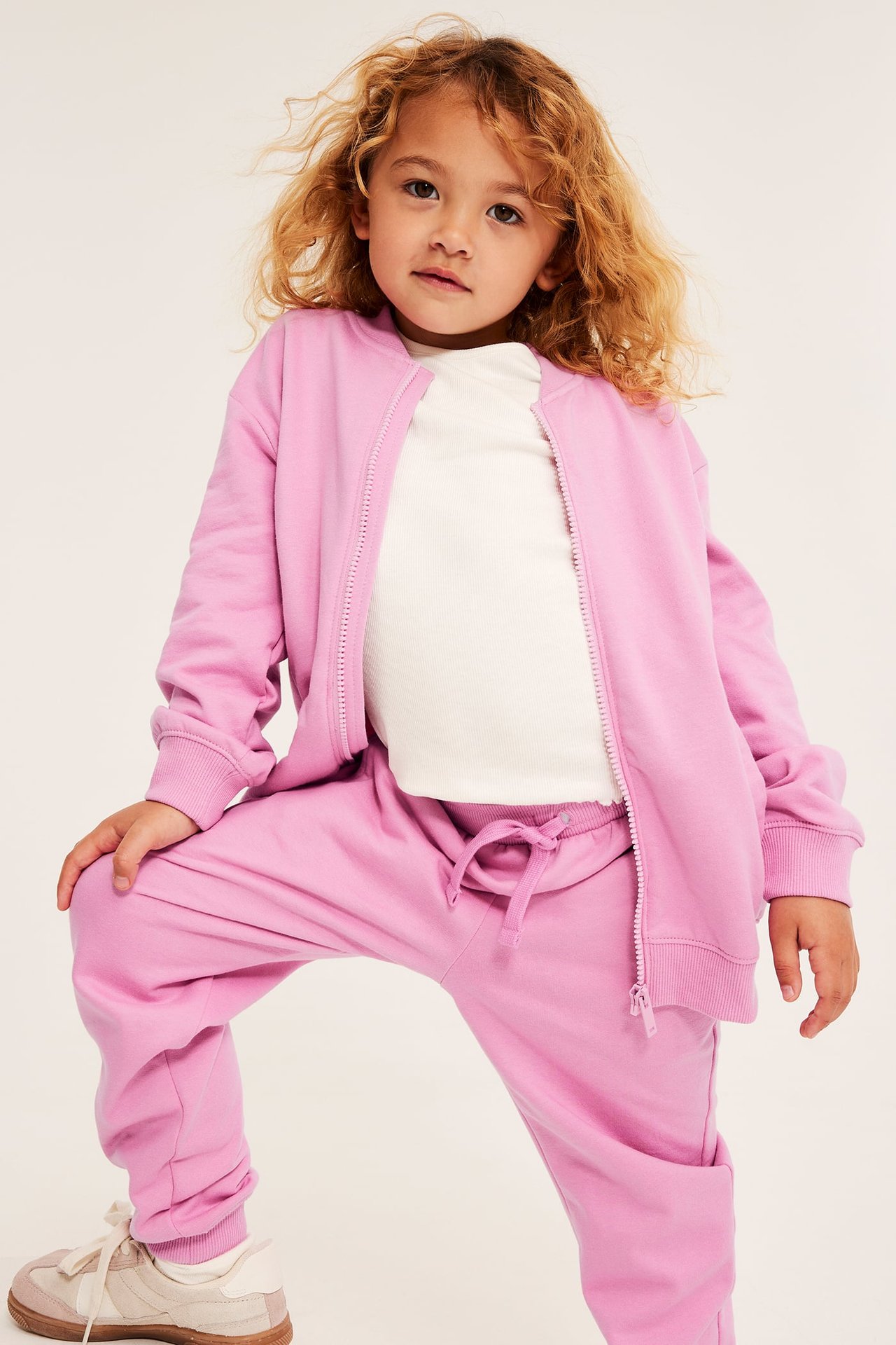 Framifrån, ett barn i rosa dragkedjetröja och mjukisbyxor, vit topp och beiga sneakers.