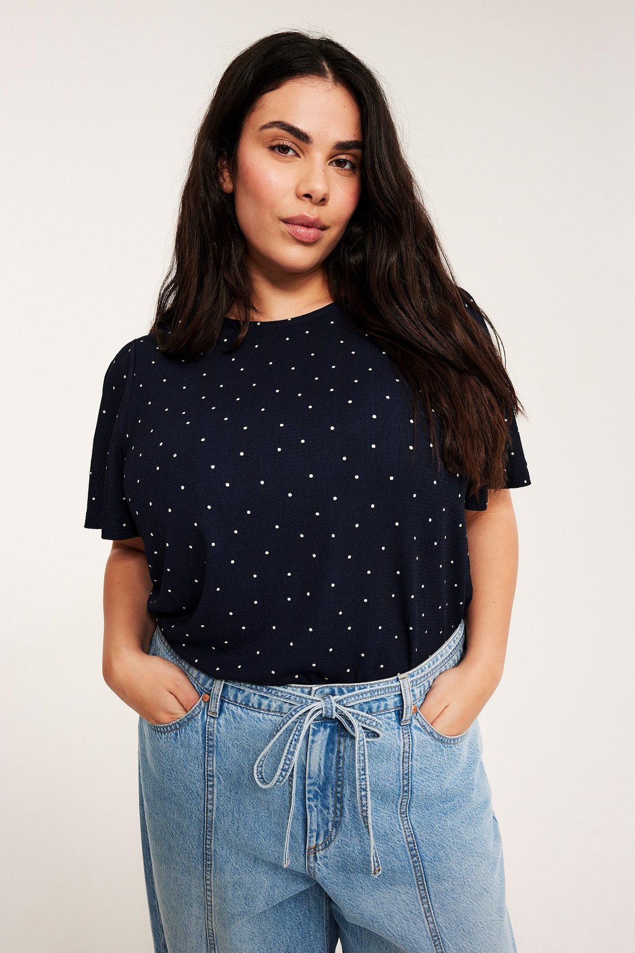 Front: Kvinne i mørkeblå prikkete kortermet topp og lyseblå jeans med snøring.