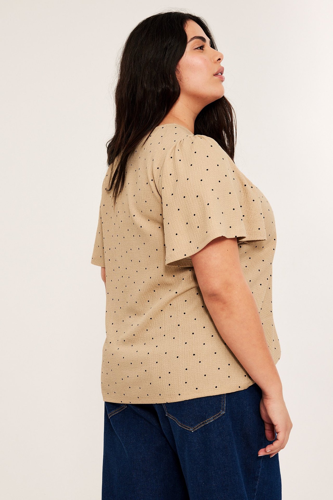 Sidebilde av en kvinne i en beige kortermet prikkete topp og mørkeblå jeans.