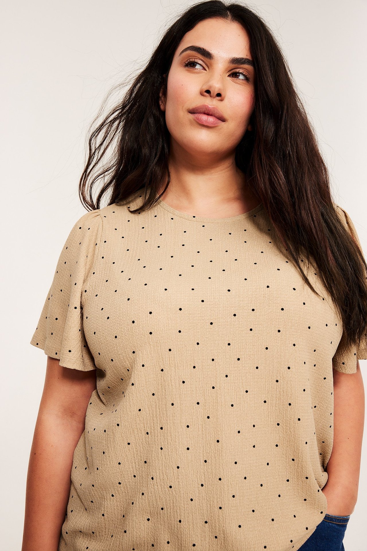 Forfra, en kvinne har på seg en beige kortermet prikkete topp med sorte prikker og blå jeans.