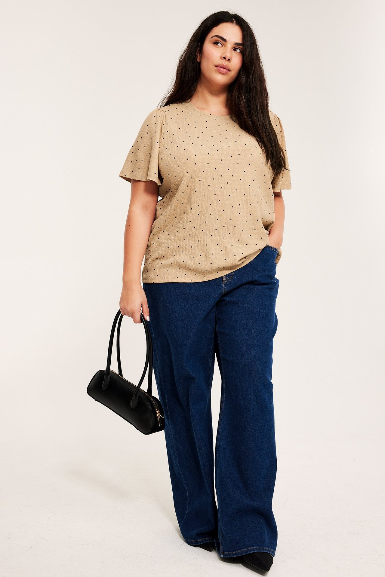 Front: kvinne i beige ribbet prikkete topp med rysjeermer, mørkeblå vide jeans, holder svart håndveske.