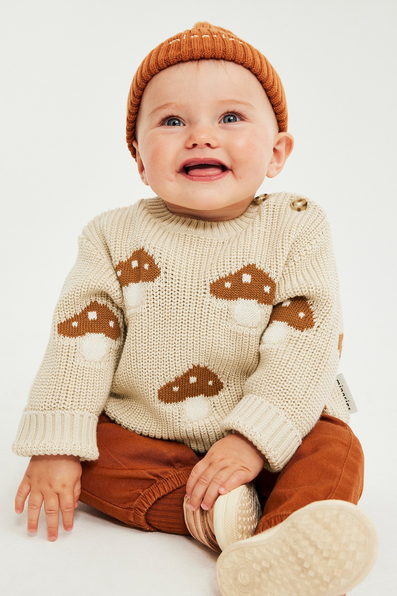 Front, baby i beige strikket genser med soppmønster, brun lue og rustfargede bukser.