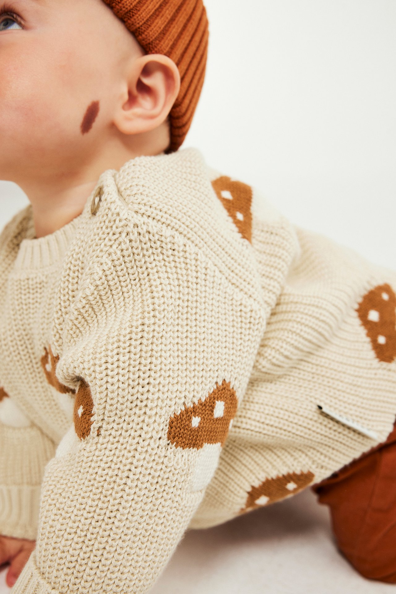 Nærbilde av baby i beige strikket genser med brunt soppmønster og rustfarget ribbestrikket lue.