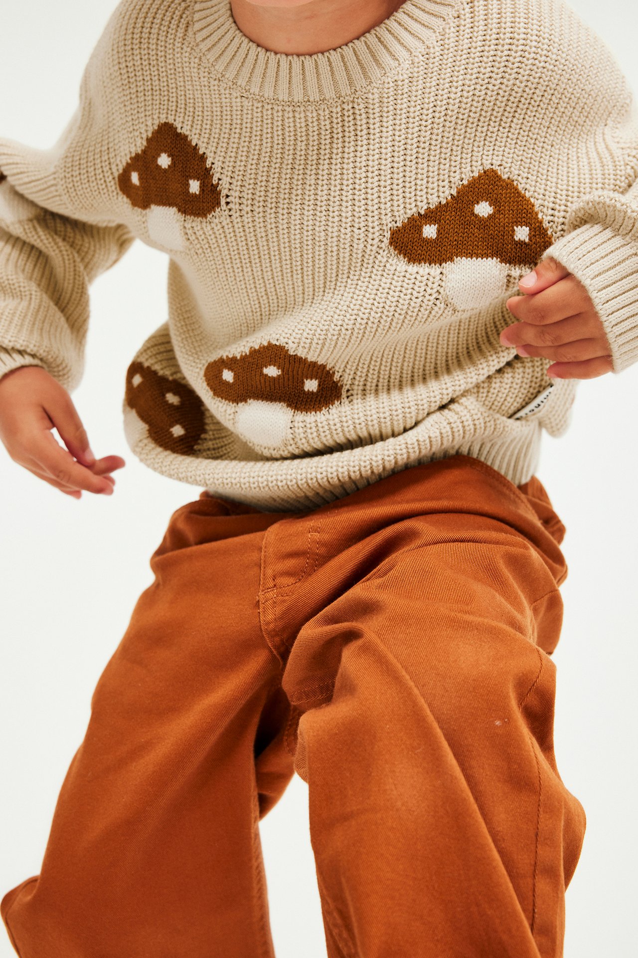 Närbild på ett barn i beige stickad tröja med svampmönster och roströda byxor.