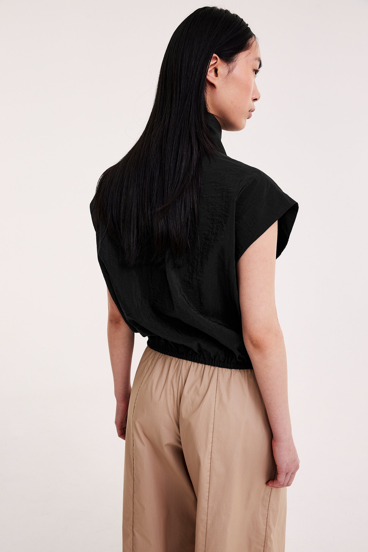 Bakifrån: Kvinna i svart ärmlös krinklad skjorta med dragsko i midjan, över beiga byxor.