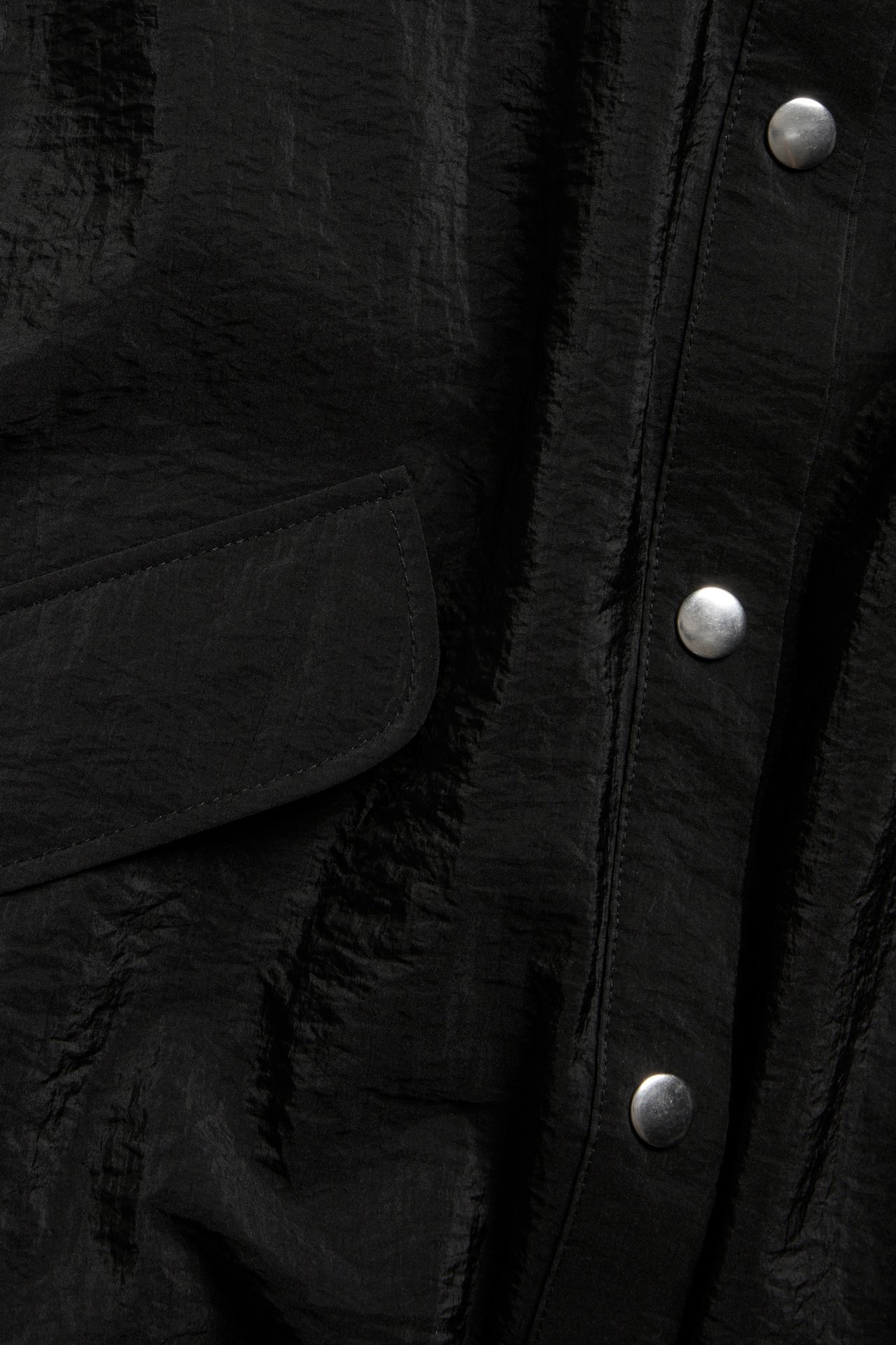 Närbild av en svart ärmlös skjorta i krinklad polyamid med ficka med lock och silverfärgade tryckknappar.