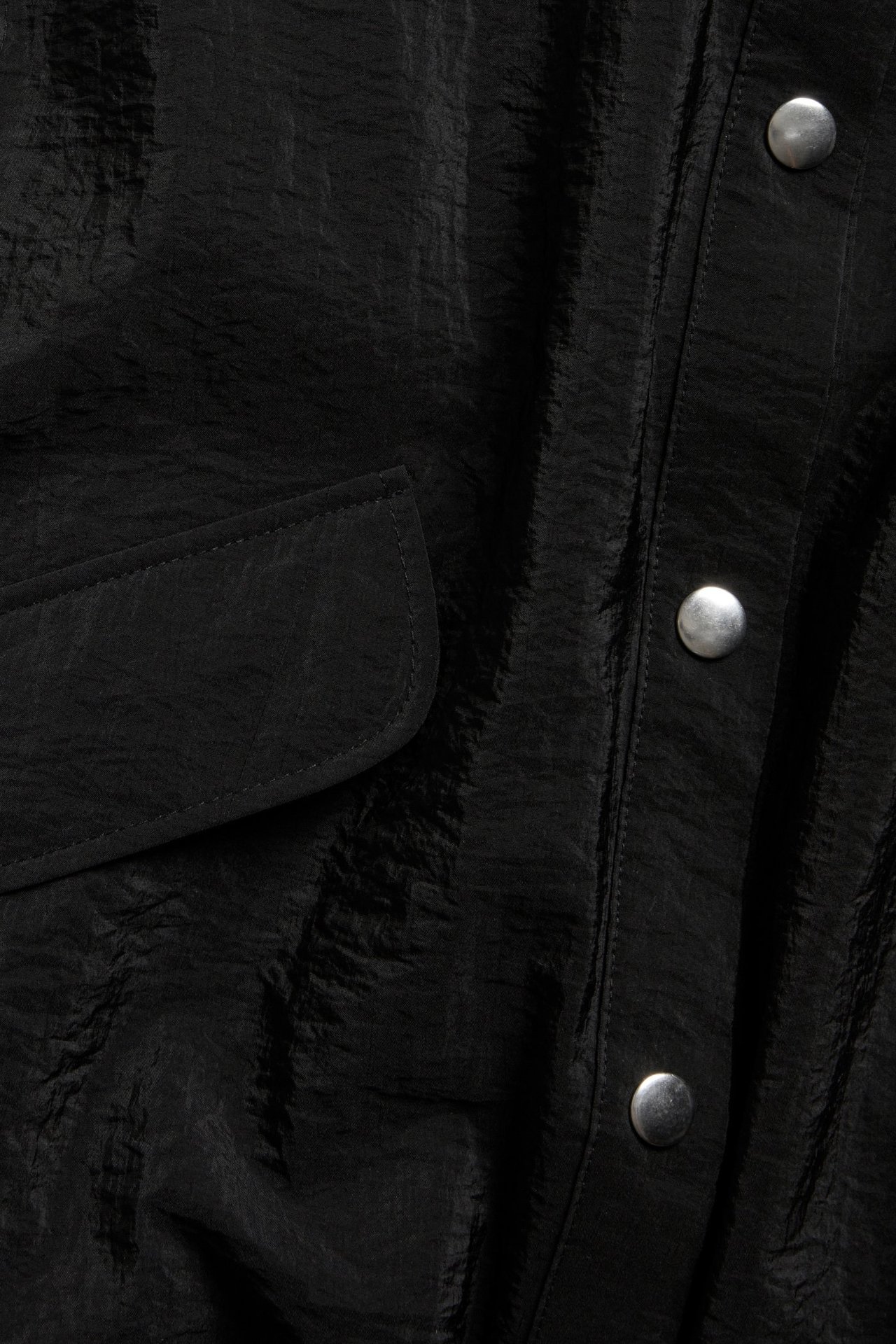 Zbliżenie czarnej, bezrękawnikowej koszuli z poliamidu, z pomarszczoną fakturą, kieszenią z klapką i srebrnymi zatrzaskami.
