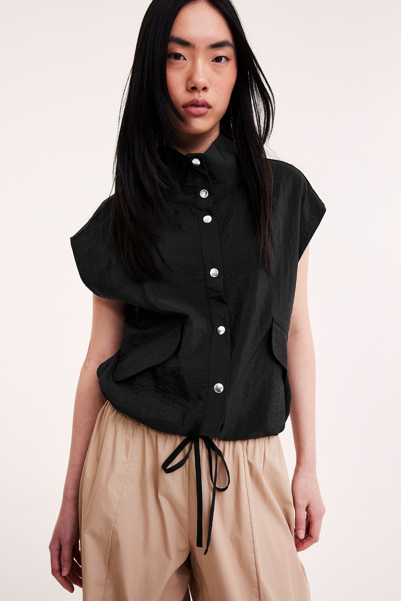 Framifrån: Kvinna i svart ärmlös skrynklig skjorta med silverknappar, dragsko i midjan, med beiga byxor.