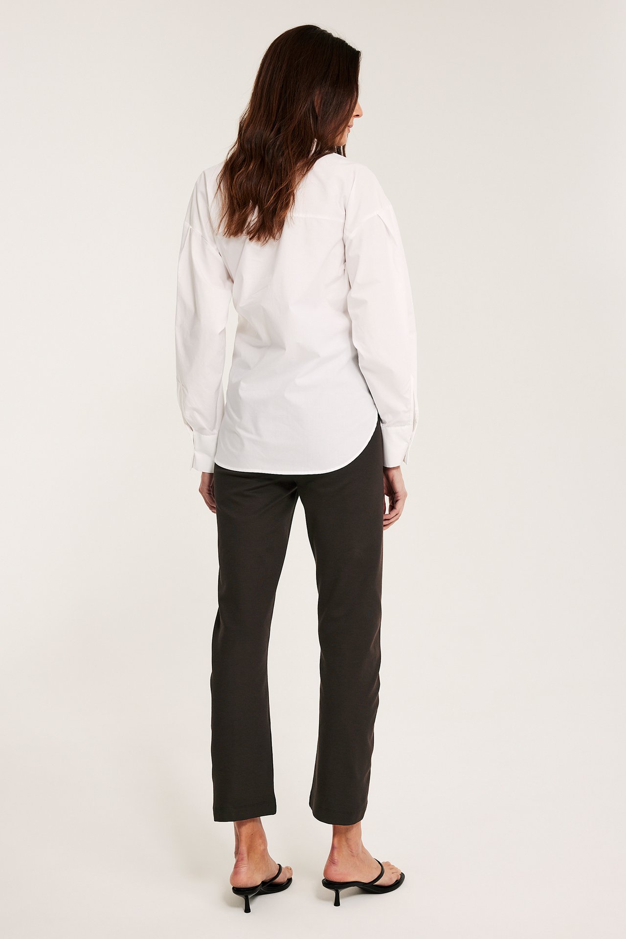 Widok z tyłu kobiety w ciemnobrązowych prostych spodniach pull-on ze szwem, białej koszuli i czarnych sandałach.