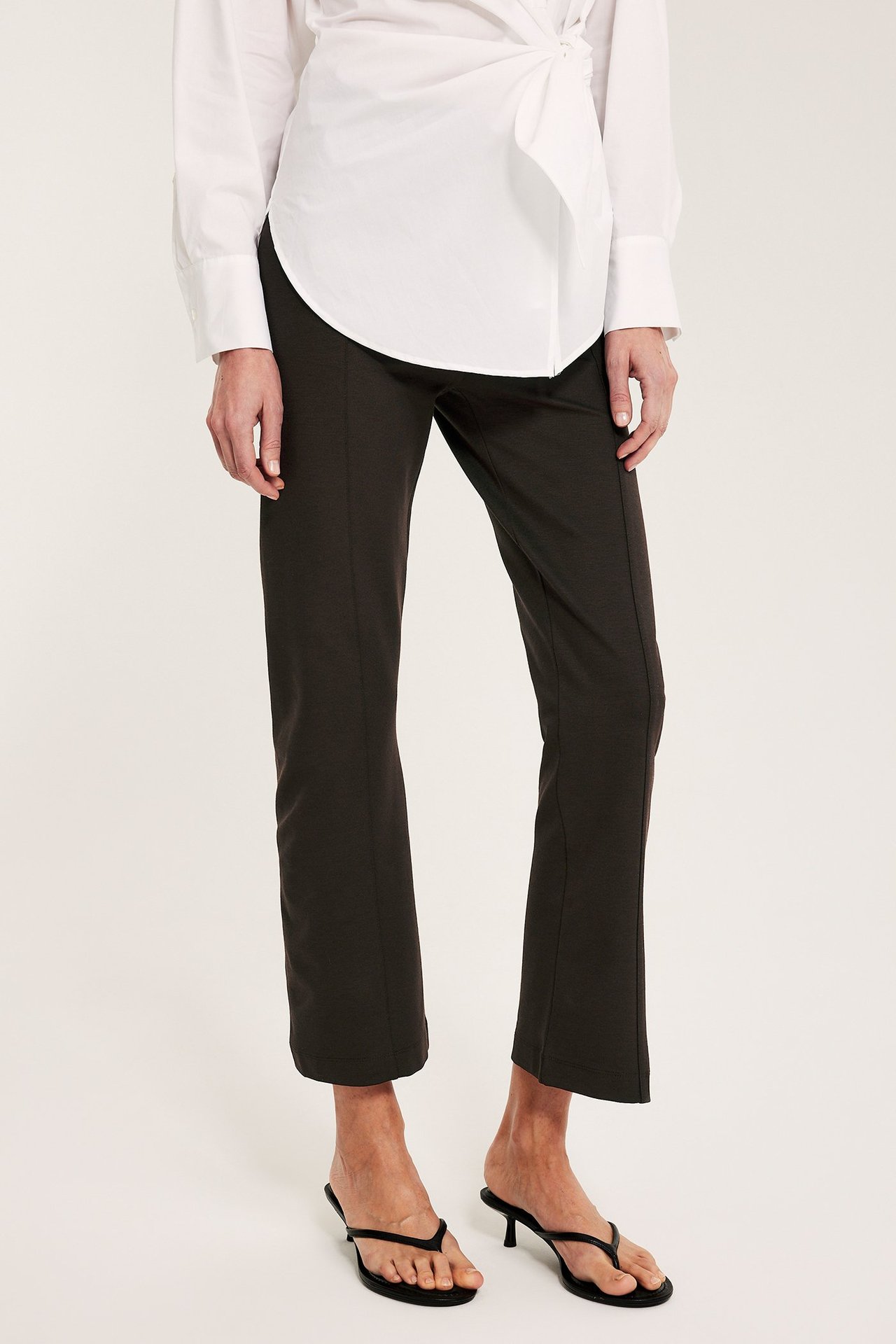 Kvinnans mörkbruna raka pull on-byxor, vit omlottsblus och svarta klackade tåsandaler.