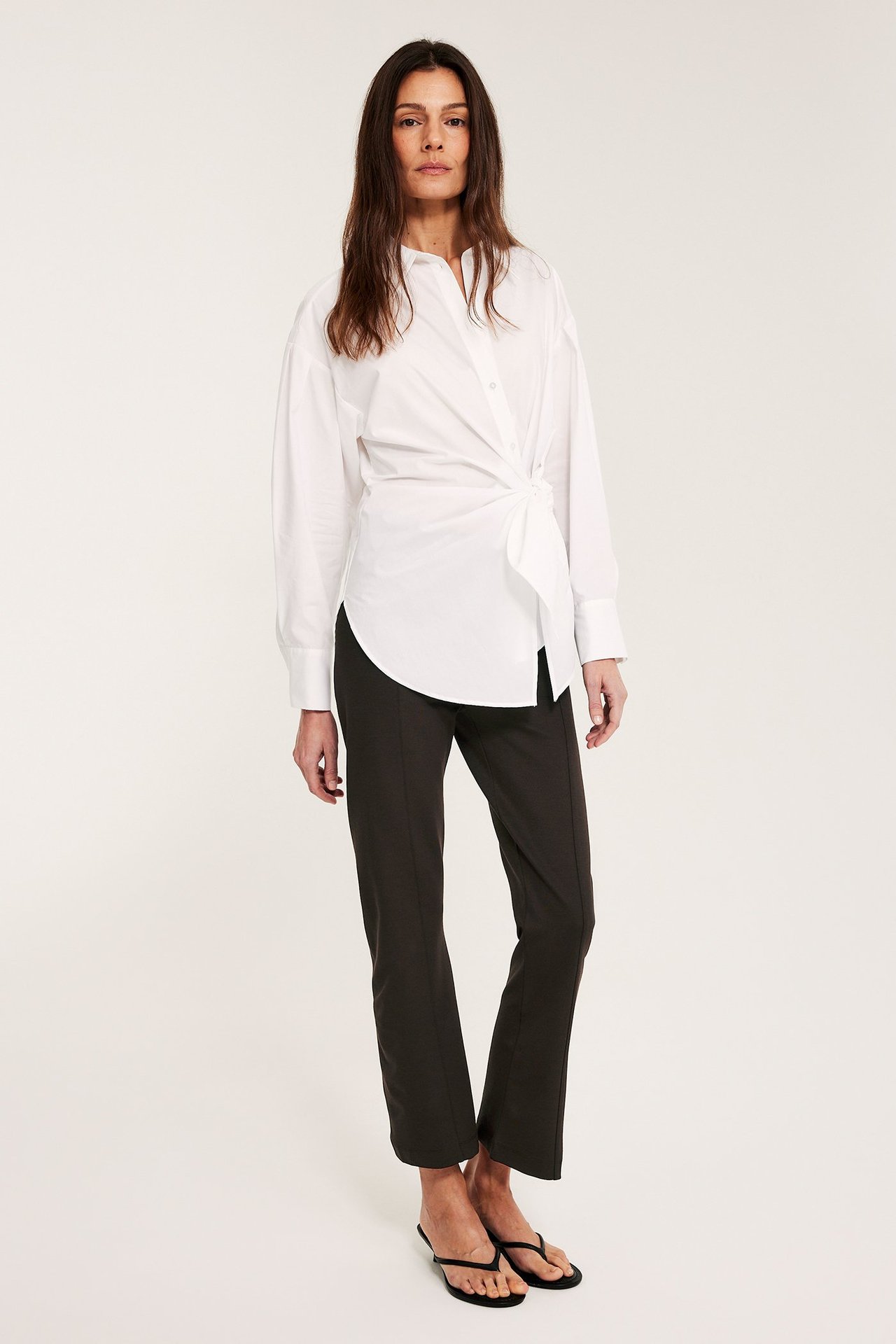 Framifrån: kvinna i mörkbruna raka pull on-byxor med söm fram, vit knuten skjorta och svarta flip-flops.